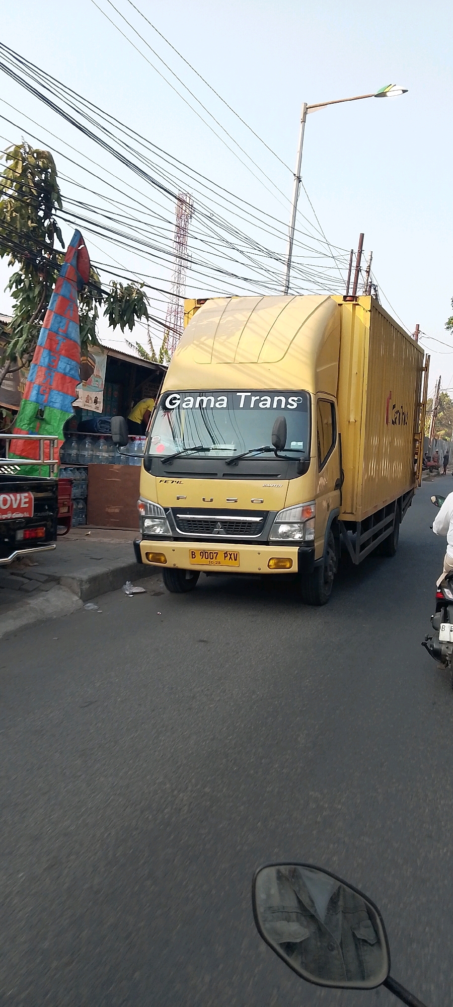 The image captures a street scene in Bekasi, Indonesia, dominated by a yellow FUSO Canter truck with "Gama Trans" branding on its windshield. The truck is parked or moving slowly on a paved road. In the background, there are buildings, trees, and a prominent cell tower. Various power lines crisscross the sky, obscuring parts of the background. To the left of the truck, there is a black pickup truck with the word "LOVE" visible on its rear. On the right side of the frame, a motorcycle is partially visible, with its rider wearing white. The truck's license plate reads "B 9007 PXV". The environment appears to be urban, with evidence of commercial activity and infrastructure.