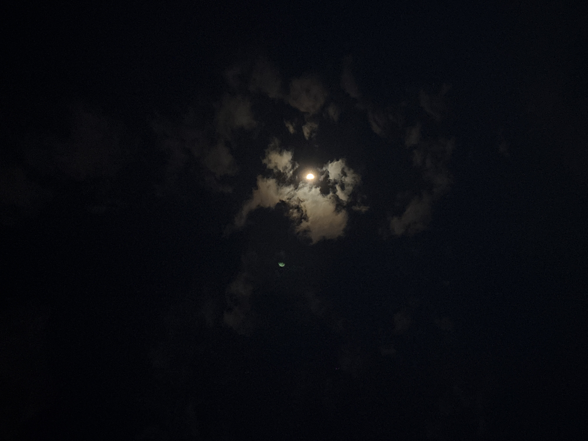 This nighttime photograph captures a celestial scene dominated by a bright moon partially obscured by clouds. The main subject is the moon, visible as a luminous, circular disk of light near the upper center of the frame, peeking through a break in the cloud cover. Its intense glow illuminates the edges of the surrounding clouds, creating halos and bright patches against the otherwise dark sky. The clouds themselves are varied, ranging from thick, dark masses to wispy, translucent formations that allow the moonlight to filter through.
A notable and intriguing detail is a smaller, crescent-shaped light source located below and slightly to the left of the main moon. This secondary object exhibits a distinct greenish hue. Its shape and color suggest it could be a lens flare or an internal reflection within the camera's optics, rather than a natural celestial body.
The overall setting is a dark, cloudy night sky. The extensive darkness in the majority of the image, coupled with the visibility of the moon and clouds, indicates an outdoor environment. There are no visible people, man-made objects, or discernible ground features. The image is characterized by deep blacks and varying shades of gray in the clouds, with the bright moon serving as the primary light source. There is no visible text in the image. The scene conveys a sense of quiet nocturnal vastness.