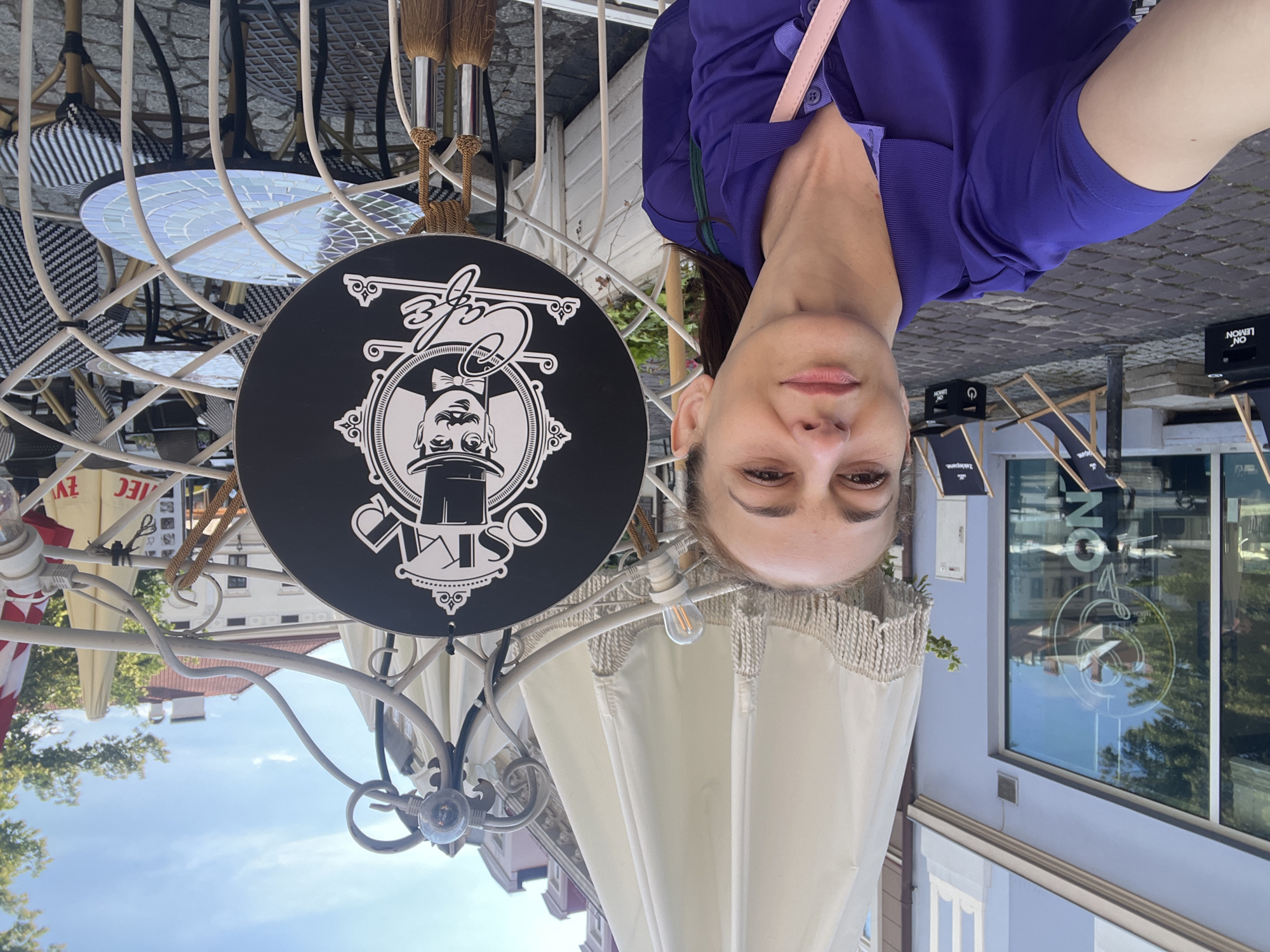 In an outdoor cafe setting, a woman with brown hair pulled back is taking a selfie. She is wearing a purple polo shirt and a pink strap from a bag is visible across her chest. To her right, a circular black sign with a white logo is hanging on a white metal frame. The logo depicts a man in a top hat with a mustache, and the text "OSKA Cafe" in cursive. Behind the sign, tables and chairs with striped cushions are set up. In the background, a building with large windows and the reflection of trees and buildings is visible. Deck chairs with the brand "ON LEMON" are also present on the cobblestone pavement. The sky above is blue with some clouds.