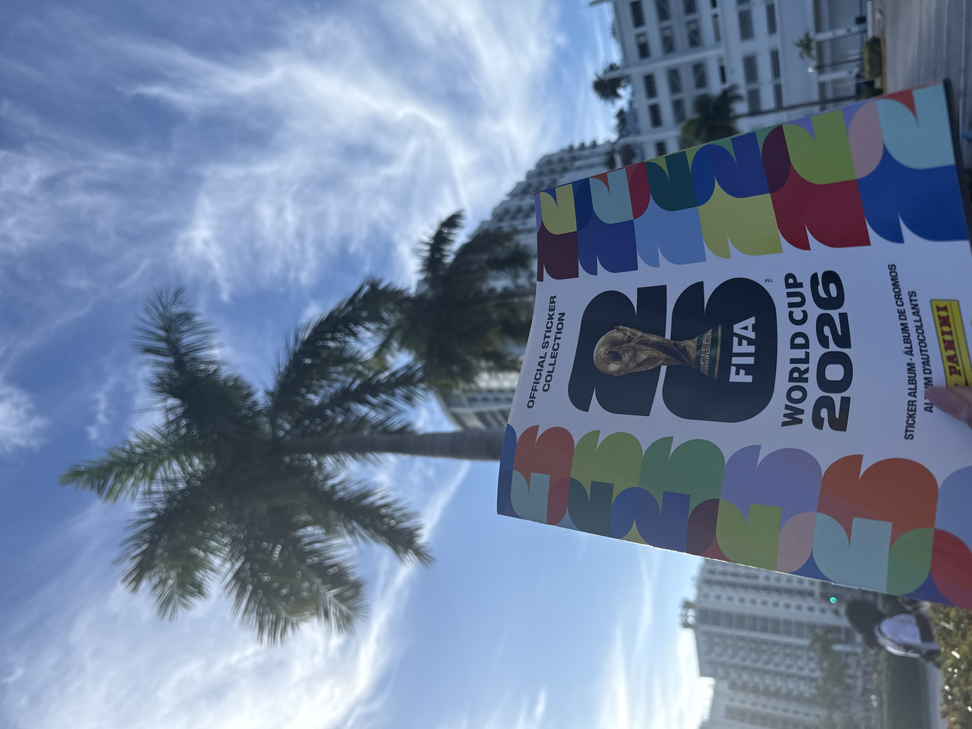 The image captures a sunny outdoor scene in Hollywood, United States of America, with a palm tree and modern buildings forming the backdrop. The primary subject is a FIFA World Cup 2026 sticker album, prominently displayed in the foreground. The album's cover features a colorful, abstract design with the FIFA logo and "World Cup 2026" text, along with the official Panini logo. The sky is blue with light, wispy clouds, providing a vibrant contrast to the urban elements.
