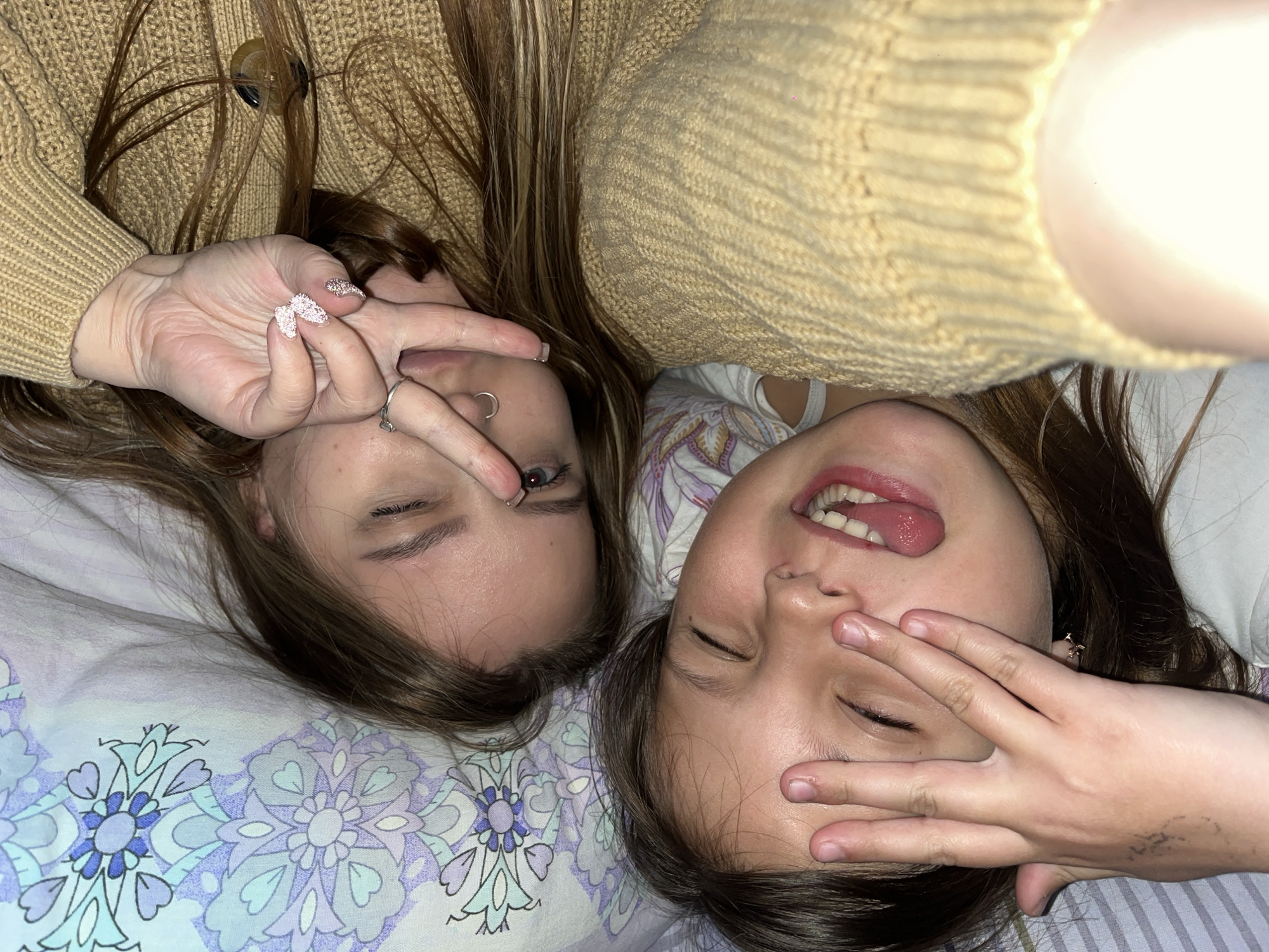Two young women are lying on a bed with a floral patterned sheet. The woman on the left is wearing a yellow sweater and making a peace sign with her hand. She has a nose ring. The woman on the right is sticking her tongue out and covering her eyes with her hand. The photo appears to be taken at close range and from a slightly rotated perspective. There is no visible indication of the city of Novopokrovskaya, Russia.