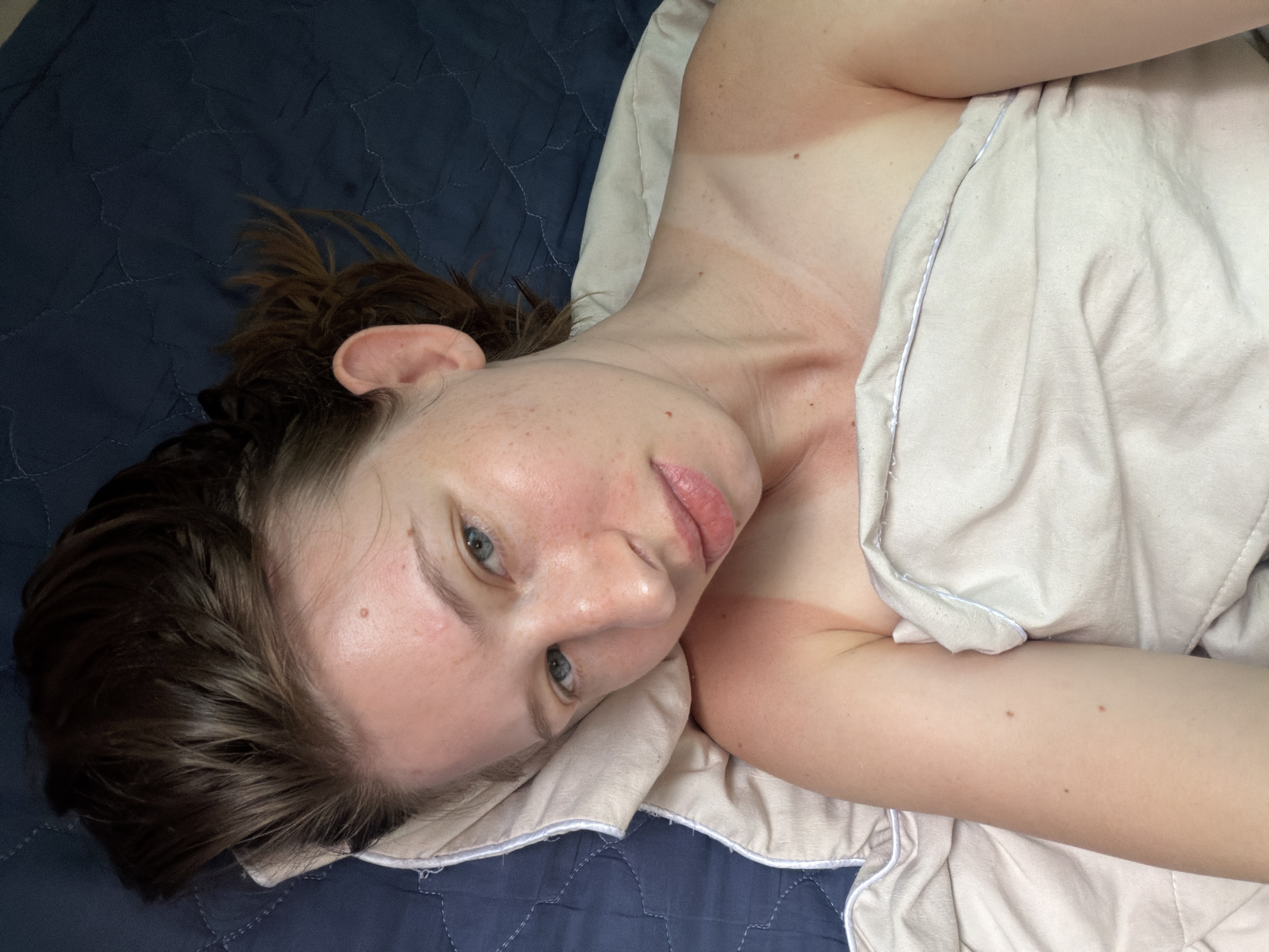 A woman lies on a blue quilted bedspread, partially covered by a cream-colored blanket. Her head is tilted back, and her face is looking towards the camera. She has brown hair, blue eyes, and her lips are slightly parted. There are visible tan lines on her neck and shoulders, suggesting recent sun exposure. The image was taken in Dongducheon-si, South Korea.