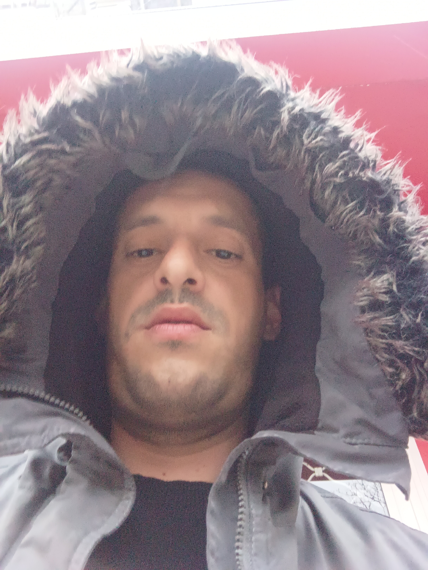 A close-up, low-angle selfie of a man with light brown skin, dark hair, and a stubbly beard. He is wearing a grey hooded jacket with a fur trim around the hood. The hood is up and obscures the background. The man is looking directly at the camera. The background is out of focus but appears to show a red surface with a white bar above it. The location is unknown and cannot be identified as Burgas, Bulgaria.