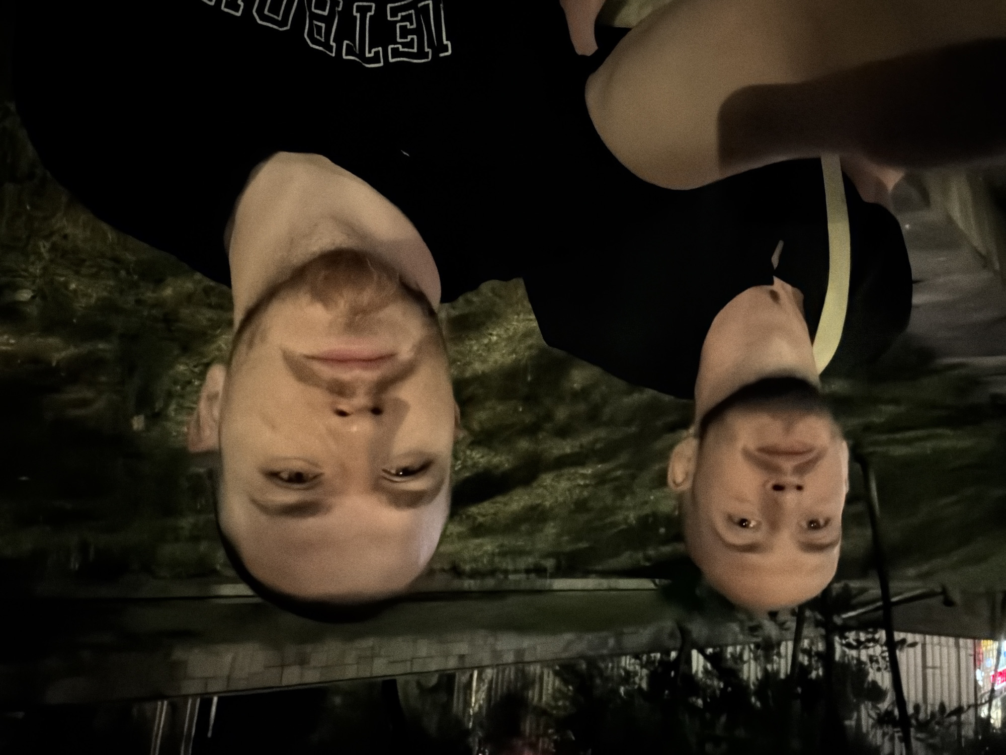 Two adult males are in the foreground of a nighttime outdoor scene, likely in a park or public square in Sofia, Bulgaria. The man on the right is closer to the camera, his face filling a significant portion of the frame. He has a shaved head, a light beard, and is wearing a black t-shirt with white lettering that appears to say "DETROIT." His gaze is directed forward, slightly to the left of the camera. The man on the left is positioned behind and to the left of the other man. He also has a shaved head and a beard, and is wearing a black t-shirt with a yellow strap across his chest. He is looking upwards and to the left. The background consists of dimly lit trees, foliage, and a paved or stone walkway or wall. The overall lighting is low, suggesting artificial illumination from streetlights or nearby buildings. The image has a slightly grainy texture.