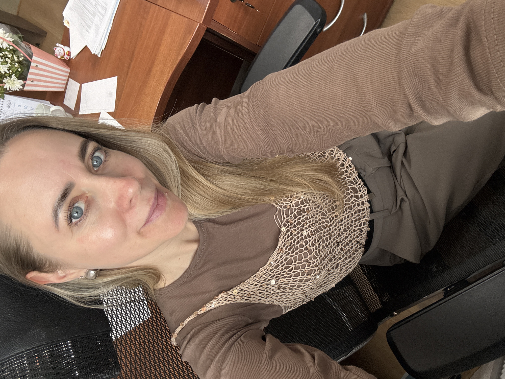 A Caucasian woman with blonde hair and blue eyes is seated in a black office chair, wearing a brown long-sleeved shirt with a sequined overlay. She is positioned near a wooden desk covered in paperwork, a bouquet of white flowers, and a striped gift bag. The image captures an overhead perspective, focusing on the woman's upper body and surrounding workspace. The location is presumed to be Chaikovsky, Russia.