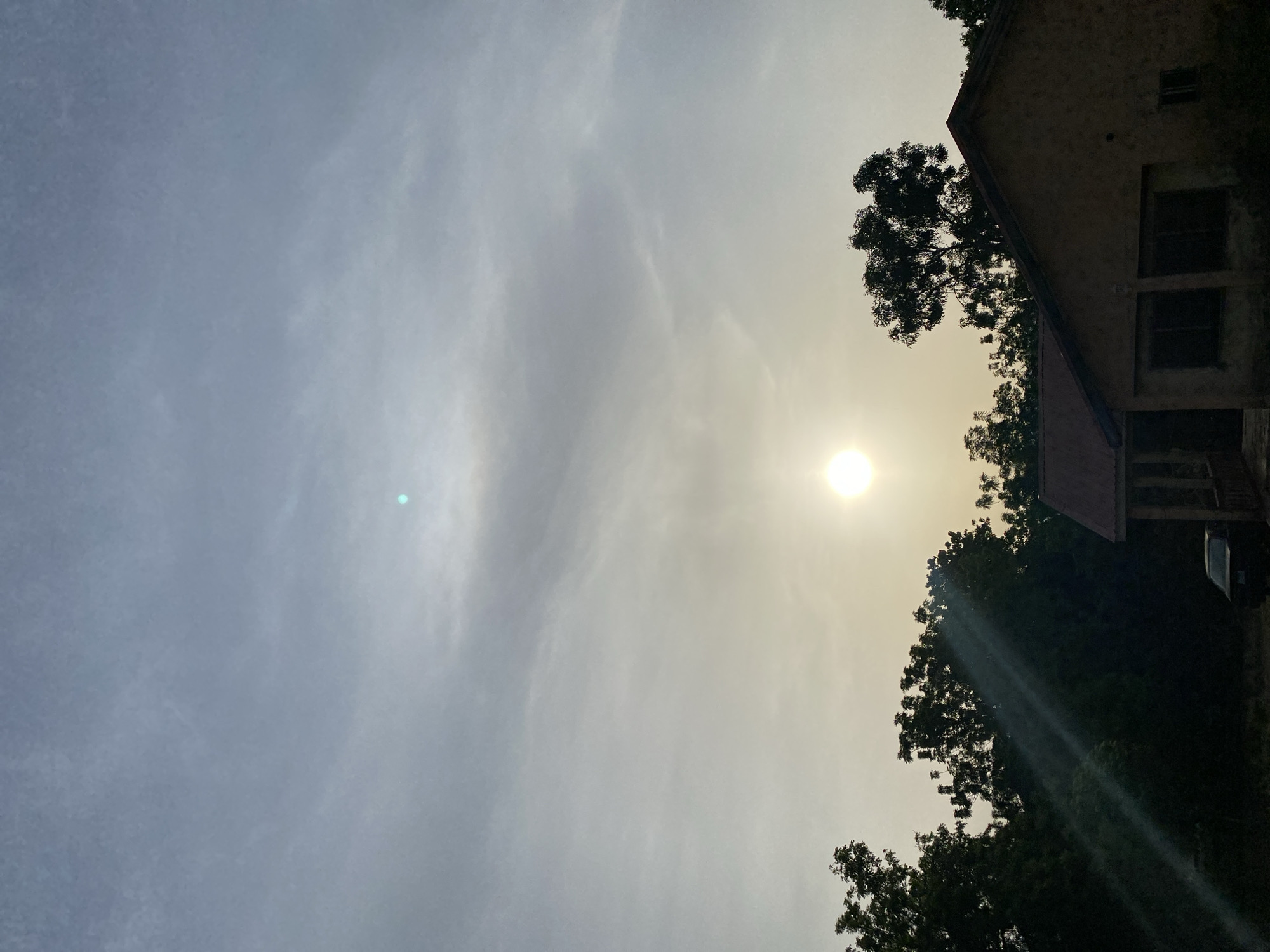 The image, taken in Adankolo, Nigeria, presents a view primarily dominated by a cloudy sky with the sun shining through. The sun is brightly visible in the upper right quadrant (when oriented correctly), appearing diffused and hazy due to thin cloud cover, and casting a warm, yellowish glow across that portion of the sky. A small, distinct teal-blue lens flare is visible in the upper left of the sky, and another elongated light streak from the sun can be seen diagonally across the lower right.
In the lower right and bottom edges of the frame, dense green trees are silhouetted against the bright sky, indicating significant backlight from the sun. Partially visible on the far right, nestled amongst the foliage, is a building, likely a house, with a discernible roofline and some window features, also appearing in deep shadow. A small portion of a vehicle is faintly visible near the building, further obscured by trees and shadows.
The lighting conditions suggest either early morning or late afternoon, given the low position of the sun relative to the foreground elements and the atmospheric haze. The weather appears overcast with high, wispy clouds. No human activity or visible text is present in the scene, which conveys a serene, natural environment with a strong interplay of light and shadow.