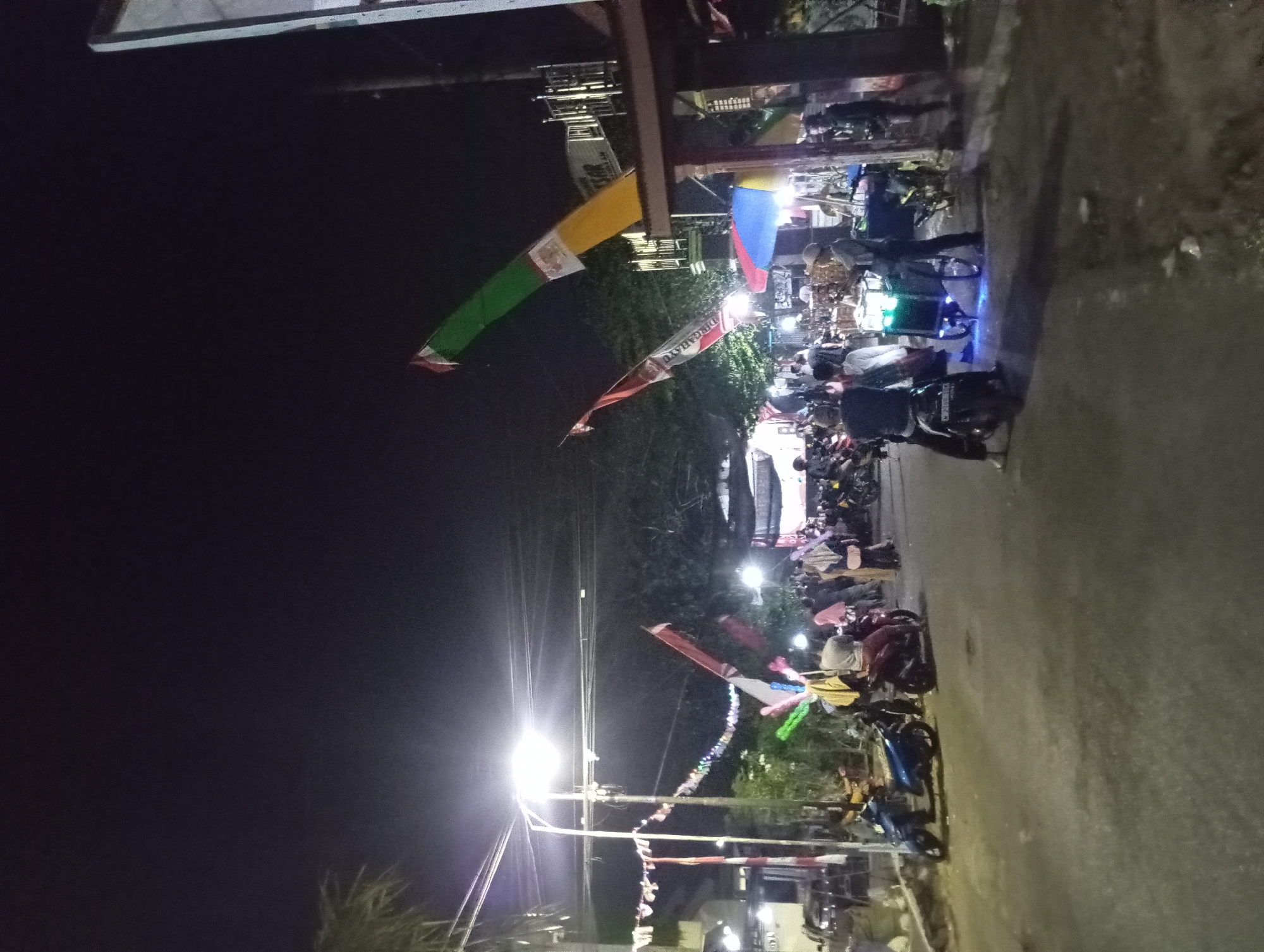 At night in Jogosari, Indonesia, a street scene unfolds with a mix of motorcycles and pedestrians. A bright street lamp illuminates the area, casting a strong light on the asphalt road and nearby trees. Red and white flags, some rectangular and others triangular, are strung across the street, possibly for a celebration. Several motorcycles are parked or being ridden, with some individuals standing near them. A food cart with glowing green lights is visible, attended by a person. In the background, there are buildings, including one with a sign that is partially obscured. The overall atmosphere suggests a lively street, perhaps a market or a gathering place at night.