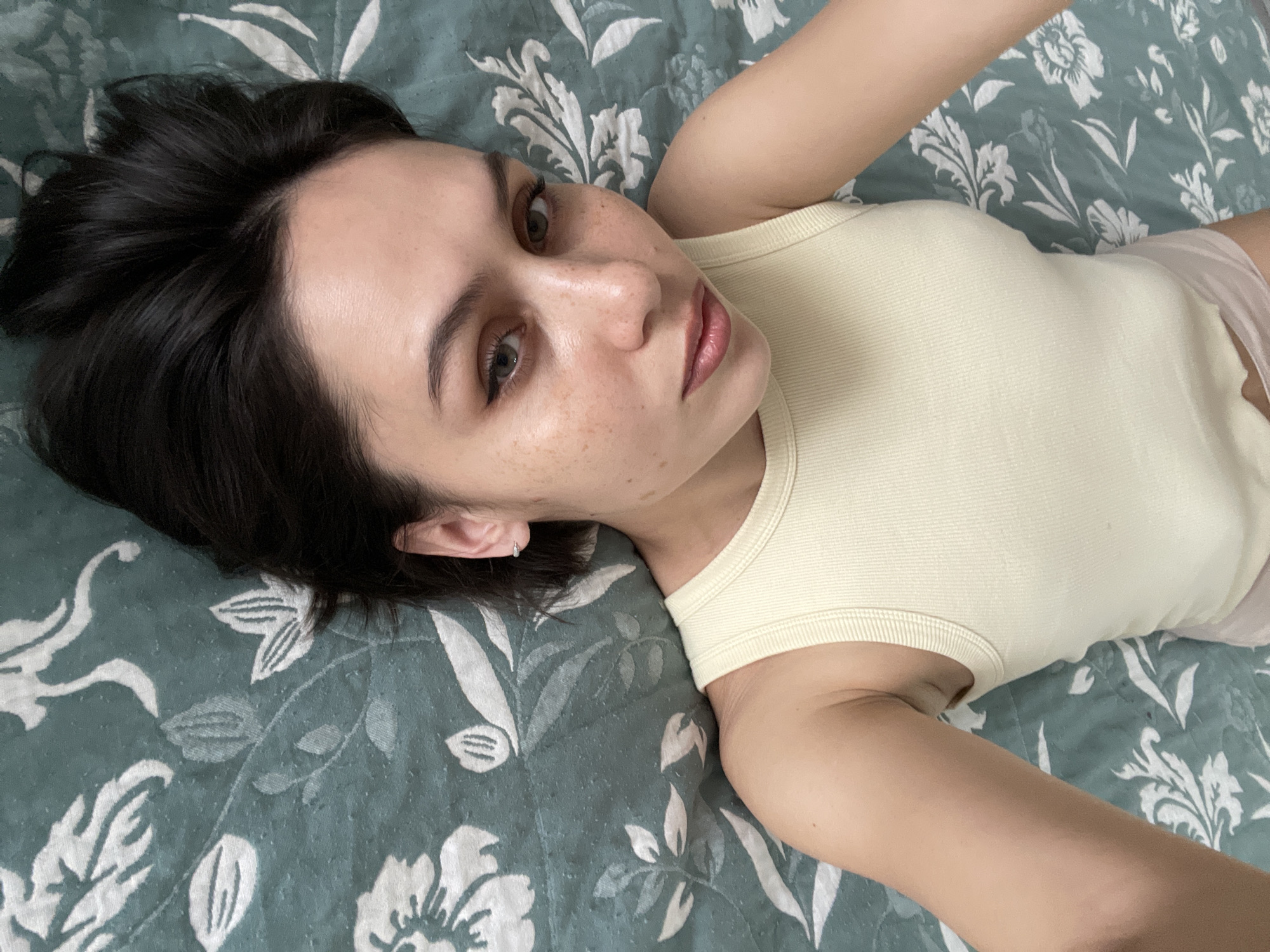 A young woman with dark hair and freckles lies on a bed with a patterned blue-green and white comforter. She is wearing a light yellow tank top. She is looking at the camera. There is no information in the image to suggest this photo was taken in Izhevsk, Russia.