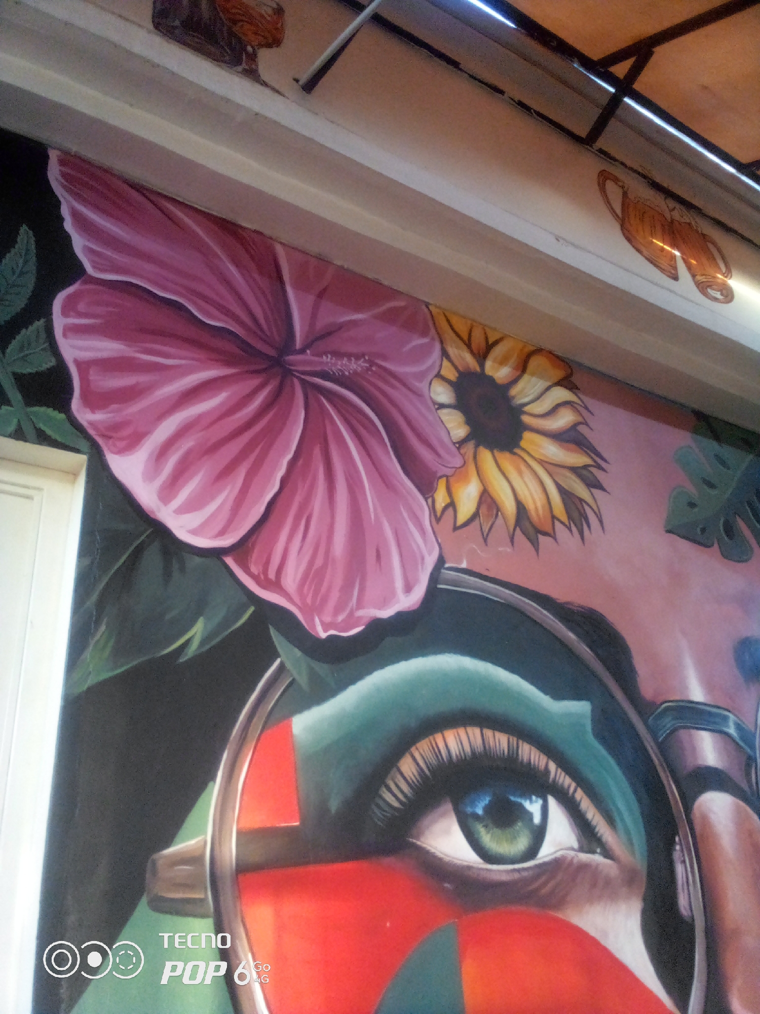 This image shows a mural painted on a wall. The mural depicts a portion of a woman's face, focusing on her eye and makeup. She has large, green eyes with long eyelashes. The makeup around her eye is a vibrant teal and red. To the left of her face, there is a large pink hibiscus flower and a sunflower. The background of the mural is a peach color with some green leaves. Above the mural, there is a white ceiling with some dark framing and what appears to be a section of a roof structure. The location of this mural is on a wall in Howayi, Nigeria. The lower left corner of the image contains a watermark with the Tecno Pop 6 Go logo.