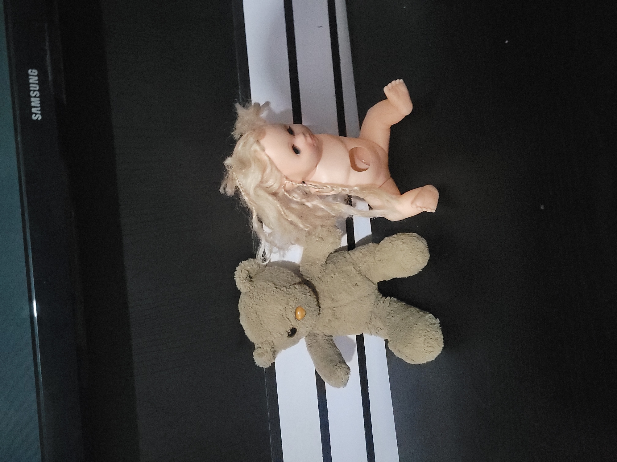 An overhead shot shows a light brown teddy bear and a vintage baby doll sitting on a black surface. The teddy bear is on the left, with its arms outstretched. The baby doll is to the right, with blonde hair and a hole in its stomach. The dolls are positioned against a backdrop of horizontal white and black stripes. In the upper right corner of the image, a Samsung logo is visible on a black surface.