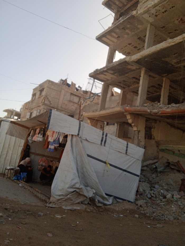 The image depicts a street scene in Khan Yunis, Palestinian Territory, characterized by significant structural damage and ongoing human activity. In the foreground, a makeshift stall or shelter constructed from light-colored tarpaulin, marked with dark stripes and yellow tape, is positioned on uneven dirt ground mixed with debris. Inside this structure, various items of clothing, including dresses and shirts of multiple colors, are suspended for display. Two individuals are present within the stall: one person, possibly an adult female, is seated on a low stool, wearing a light head covering and dark clothing, looking downwards; another person, dressed in dark clothing, is crouched lower to the ground, partially obscured.
To the right of the stall, a multi-story reinforced concrete building exhibits severe structural damage, with multiple floors partially collapsed or fragmented, exposing concrete columns, fractured floor slabs, and reinforcement bars. Piles of concrete rubble and general building debris accumulate at its base. In the background, another multi-story building, constructed of lighter-colored masonry, also shows considerable damage, including a compromised roof and broken windows. Overhead, thin utility wires traverse the upper left portion of the frame against a clear, pale blue sky, indicating daylight. The overall environment suggests an area impacted by extensive destruction, where daily life continues amidst the wreckage.