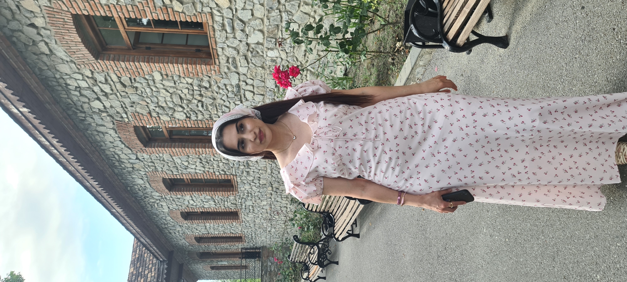 The image depicts a young woman standing in front of a stone building with a wooden roofline in Shaki, Azerbaijan. The building features arched windows with wooden frames and is constructed from large, irregular stones. Several wooden benches are positioned in front of the building, and a path runs alongside them. The woman is wearing a light pink, floral-print dress and a white headscarf. She is holding a dark-colored mobile phone in her left hand and has jewelry on her wrist and a ring on her finger. A bush with bright pink roses is visible to her right. The overall setting suggests an outdoor location, possibly a historical site or a park in Shaki.