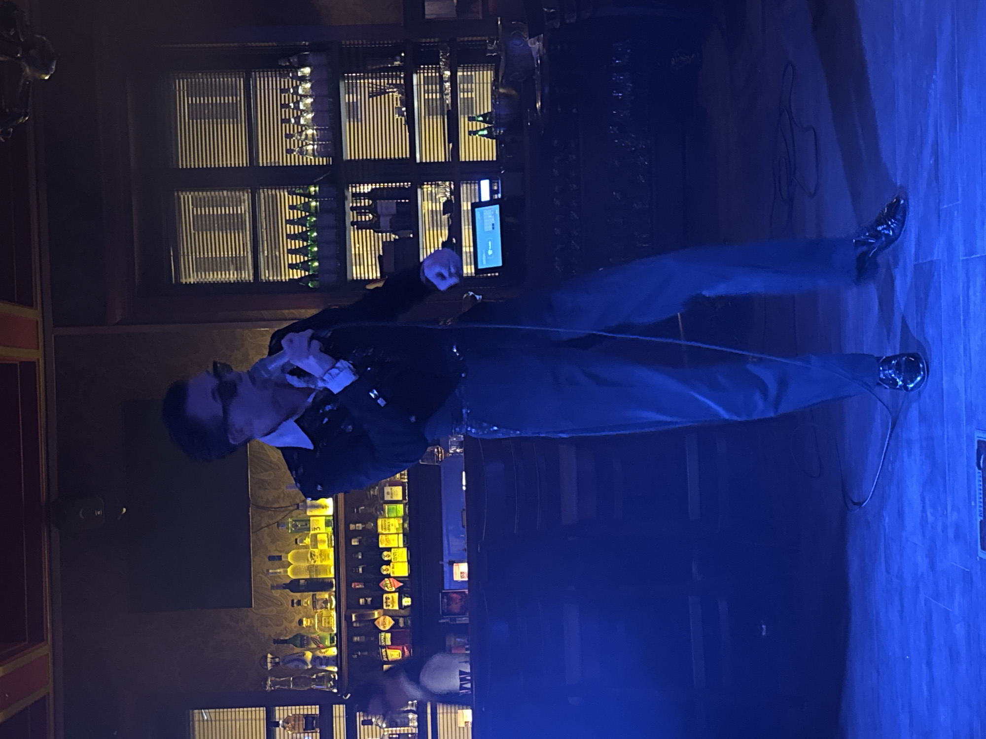 A man in a dark jacket and dark pants is singing into a microphone on a stage in what appears to be a bar. Behind him is a well-stocked bar with shelves of liquor bottles illuminated with golden light, and a window with horizontal blinds. The stage floor is blue.