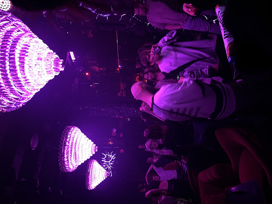 The image depicts the interior of a dimly lit establishment, likely a nightclub or lounge, in Moscow, Russia. The dominant lighting is purple/magenta, primarily originating from three prominent, tiered crystal chandeliers suspended from a dark ceiling. The walls are dark, with sections exhibiting a reflective or textured quality resembling fractured light patterns. Red upholstered armchairs or lounge seating are visible in the left foreground. An illuminated sign displaying the text "MTOO" is visible on the rear wall.
Approximately 15-20 individuals, primarily young adults, populate the space. In the mid-ground, a bald male with a beard is seen from behind, wearing a light-colored, long-sleeved shirt with dark graphics. To his right, a young male wears a light-colored, long-sleeved shirt with dark accents and text (possibly "CASHMERE") and is actively drinking from a clear glass or bottle. Further right, a male in a light grey t-shirt and jeans stands, partially obscured. A woman with long blonde hair is visible in the mid-ground left, appearing to wear a dark strip covering her eyes. People are observed standing, socializing, and consuming beverages in a crowded, social environment.
