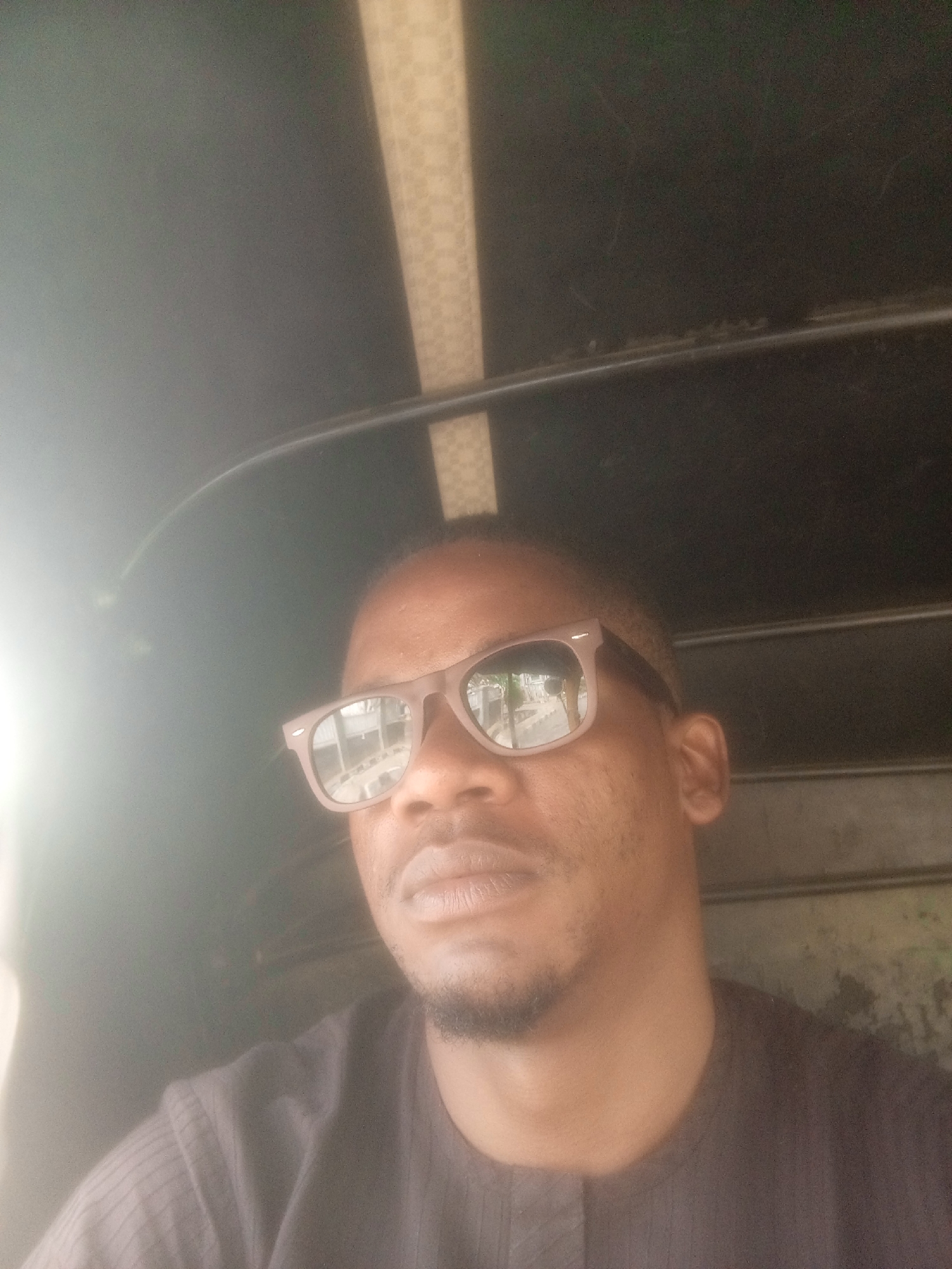 A close-up photo shows a man wearing sunglasses inside what appears to be a vehicle with a black interior. The sunglasses reflect buildings and foliage, suggesting an urban environment. The checkered pattern on the vehicle's ceiling is visible. Given the request for the location in Gwarinpa, Nigeria, one could infer he is in a commercial tricycle, commonly known as a "keke", although this isn't directly visible. The man has short hair and a small amount of facial hair. He wears a dark shirt. The lighting is partially obscured by glare.