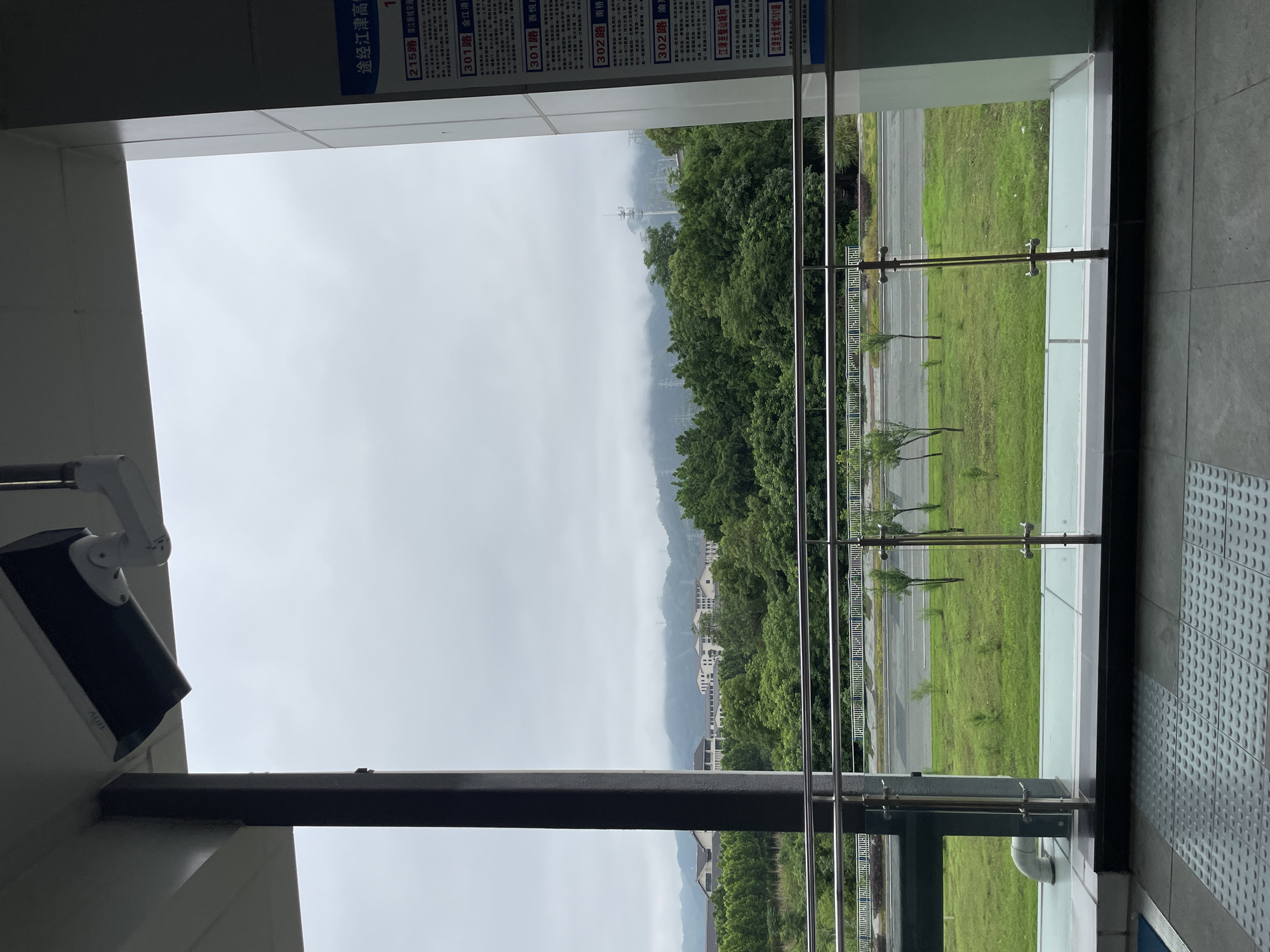 A view of Jiangjin, China, on an overcast day, seen through a window or opening. In the foreground, structural elements, including a black support bar and a white security camera, are visible. Part of a sign containing Chinese characters can be seen at the top of the frame. The view from the window reveals green trees, grass, and structures in the distance, with misty mountains in the background. Metal railings stand outside. Part of a textured gray walkway is visible on the right. The printed words "途经江津高" and the routes "215路 流通", "301路 金江港", "301路 吾悦", "302路 南桥", "302路 渝", "「江津至璧山城际", "江津至大学城C101路", and "第一大学" are readable from the sign.