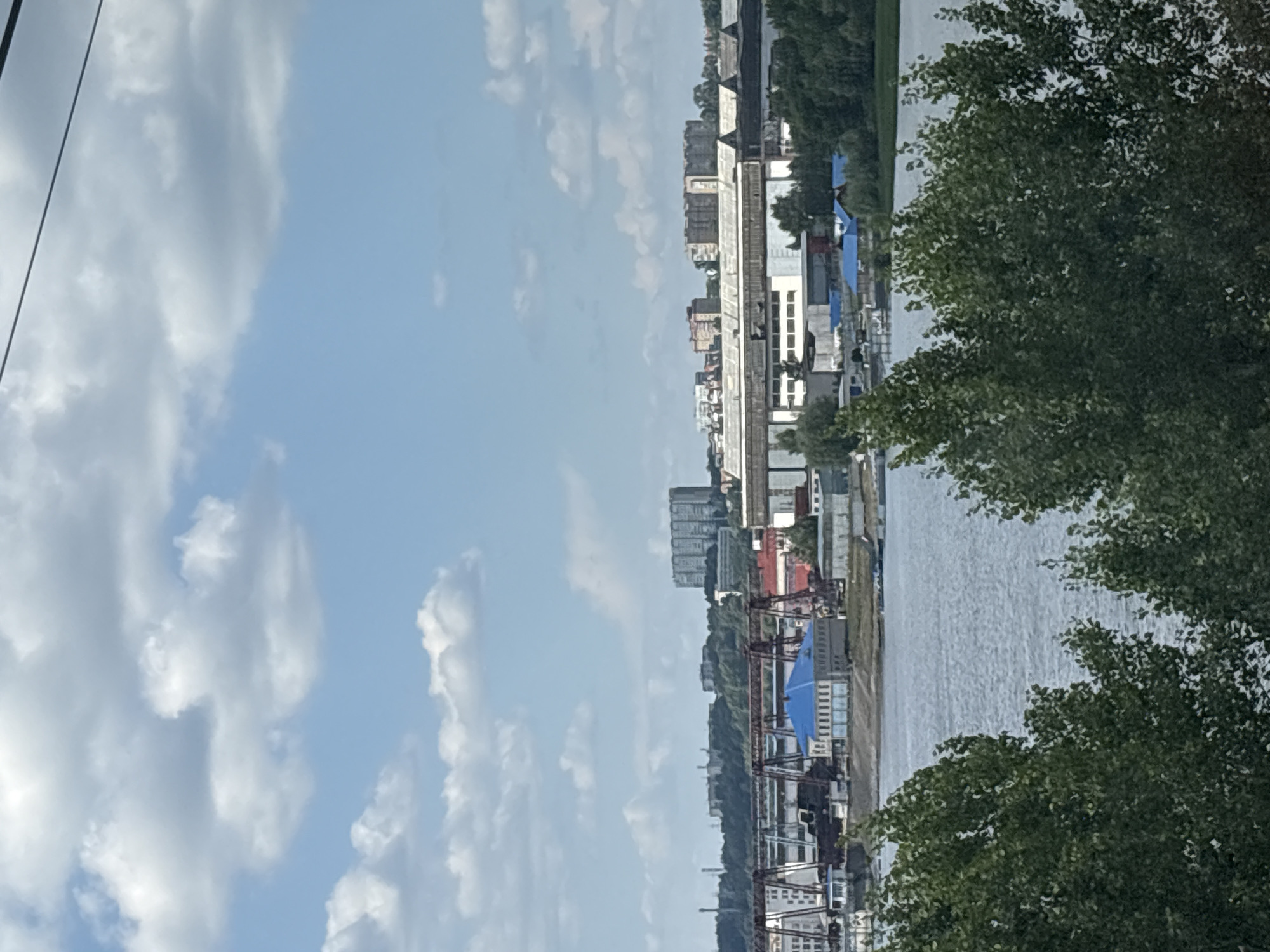 The image shows an industrial area along a body of water, likely the Volga River, in Zelenodol'sk, Russia. In the foreground, lush green trees partially obscure the view. Across the water, a complex of industrial buildings and structures is visible. These include large, long, low-rise factory-like buildings with light-colored roofs, some with visible windows or vents. Among these are several smaller buildings with distinctive bright blue roofs. In the background, a large, multi-story, block-like building complex stands out, suggesting residential or office structures. Several large gantry cranes are present in the middle ground, indicating port or industrial activity. The sky is a light blue with scattered white cumulus clouds. The overall scene depicts a waterfront industrial zone with evidence of manufacturing and infrastructure.