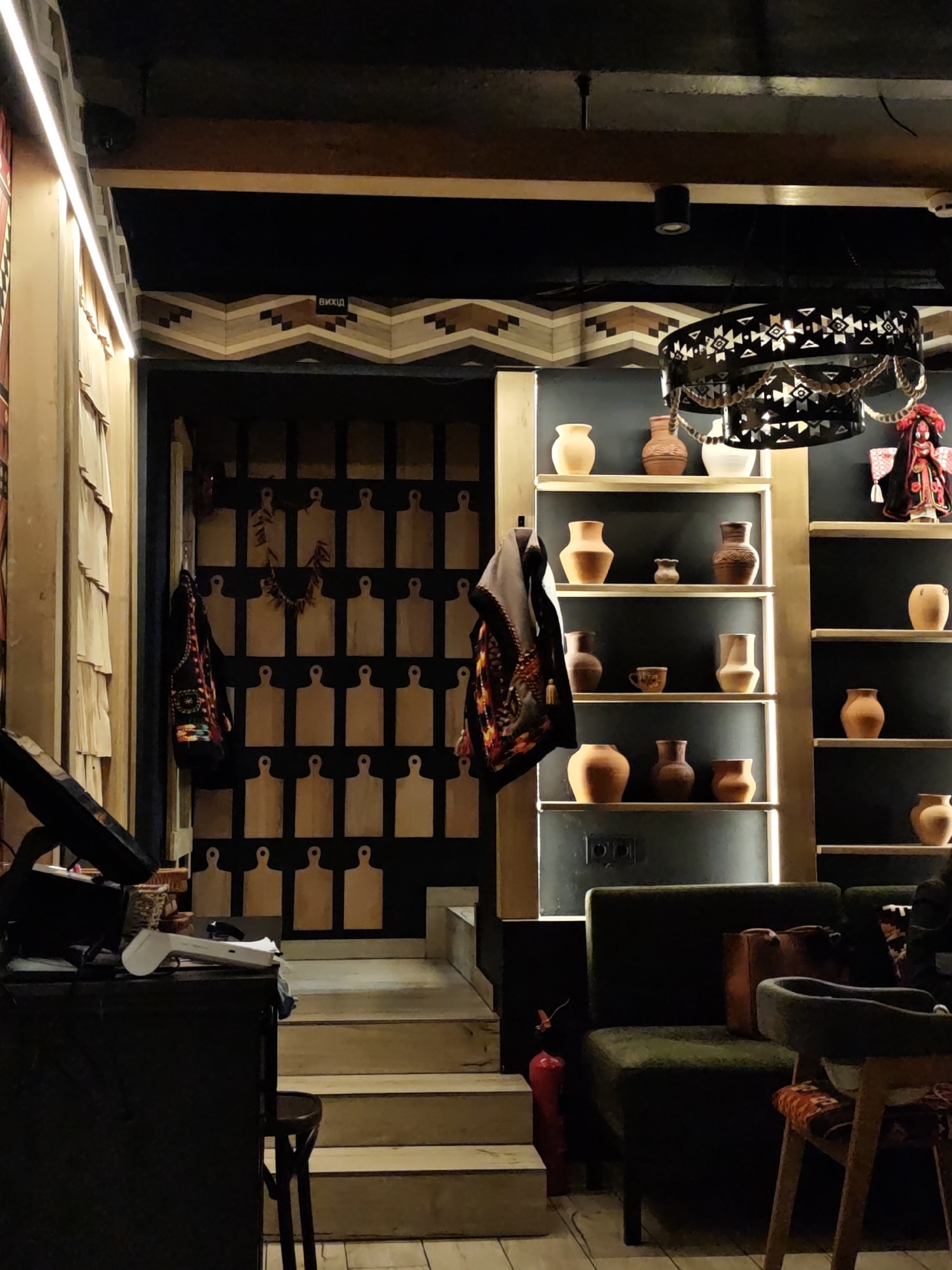 The image depicts the interior of a restaurant, likely in Ivano-Frankivsk, Ukraine, characterized by its distinctive decor. The background features a wall decorated with numerous light-colored cutting boards arranged in a grid pattern. Adjacent to this wall is a shelf unit with recessed lighting, displaying a collection of ceramic pots and vases of various shapes and sizes. A decorative chandelier with ethnic patterns hangs above. To the left, a cashier's desk is visible with a monitor and point-of-sale system. In the foreground, an olive-green upholstered chair is situated next to a wooden chair with patterned upholstery. A red fire extinguisher is located near the chair. The scene is dimly lit, contributing to a cozy ambiance. A sign with the inscription "ВИХІД" (Exit) is also visible.