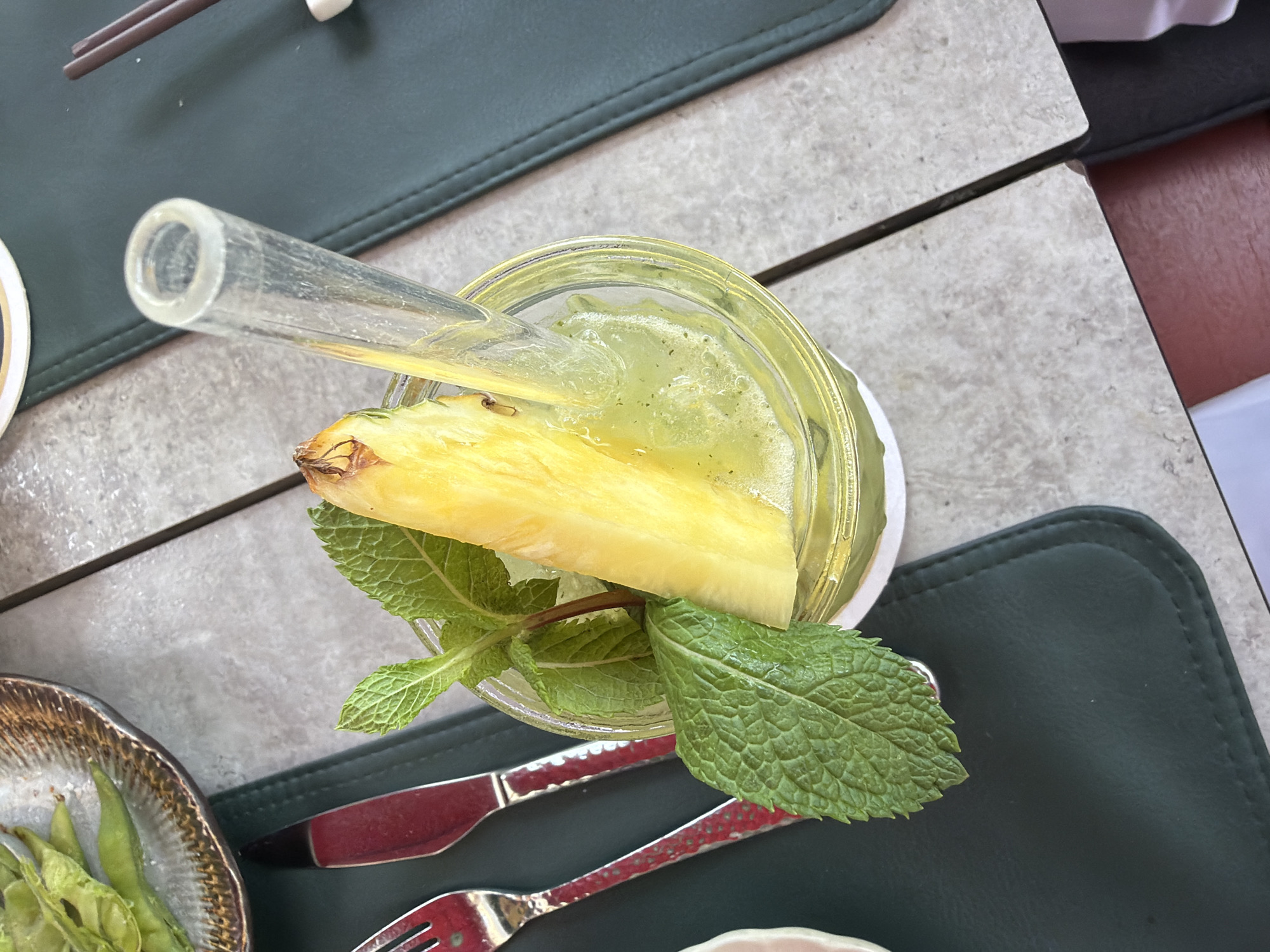 The image shows a close-up, overhead view of a refreshing green beverage in a clear plastic cup, garnished with a slice of pineapple and several sprigs of mint. A clear glass straw is inserted into the drink. The cup sits on a light-colored, tiled surface, likely a table, with a dark green placemat partially visible beneath it. To the left of the drink, a fork and knife with metallic handles are laid out on the placemat. In the upper left corner, a small dish holds edamame. In the background, chopsticks rest on a dark green surface. The overall scene suggests a meal or refreshment at an outdoor or casual dining setting. The image does not contain any identifiable landmarks or clues to pinpoint its specific location within Munich, Germany.