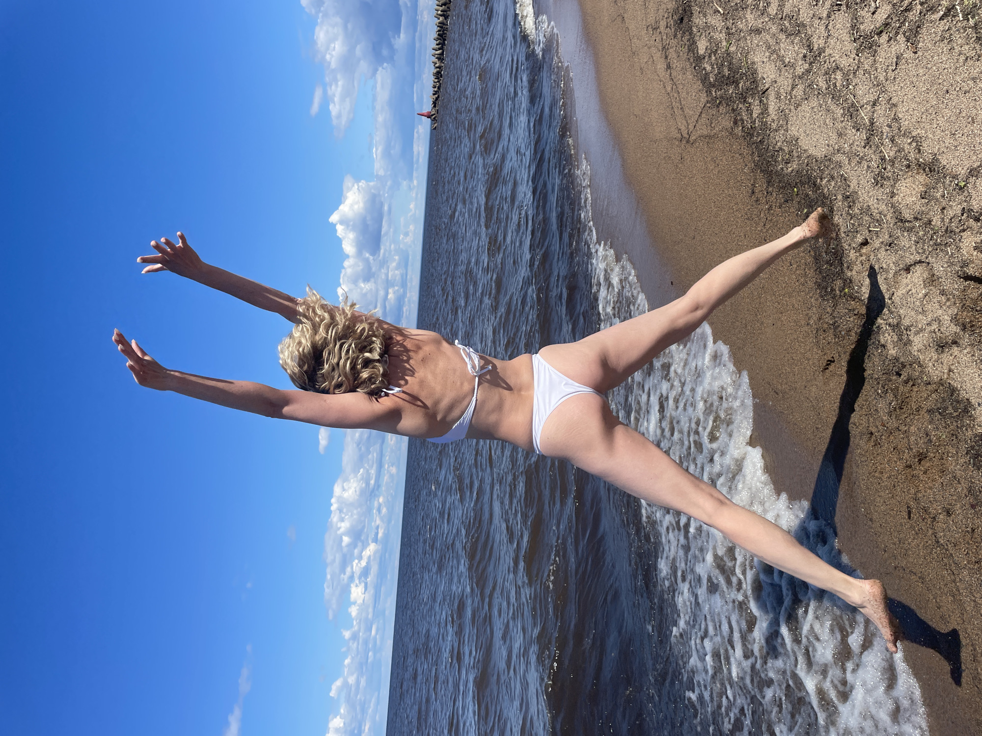 A woman in a white bikini is captured mid-jump on a sandy beach, her arms extended overhead. The sea, with small waves and a gentle surf, laps at the shore. In the distance, a breakwater with a red marker is visible. The sky is a clear blue with scattered white clouds. The scene appears to be located on the shore of Lake Beloye in the city of Belozersk, Russia, given the distinctive appearance of the lake and the presence of the breakwater, a common feature in the region.