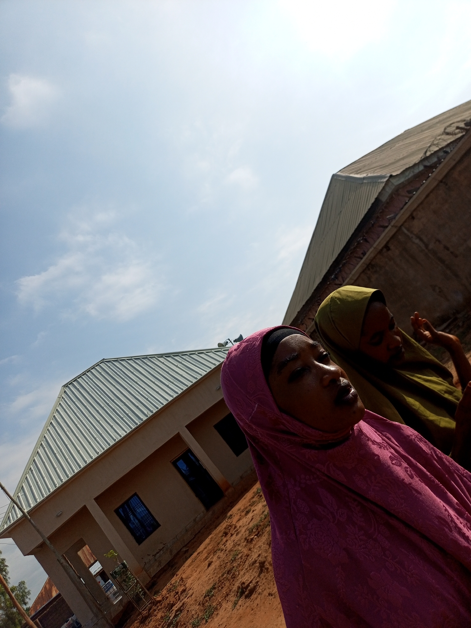 The image captures two women, adorned in hijabs, standing outdoors. The woman in the foreground wears a bright pink hijab with a textured pattern, and her face is illuminated by the sunlight. Behind her, another woman is visible, partially obscured, wearing a muted green hijab. They are positioned in front of a newly constructed building with a corrugated metal roof. The building appears to be located in a dusty, undeveloped area, with exposed soil and sparse vegetation. The sky is a clear blue with a few scattered clouds. The overall scene suggests a warm, sunny day in an outdoor setting within Gombe, Nigeria.