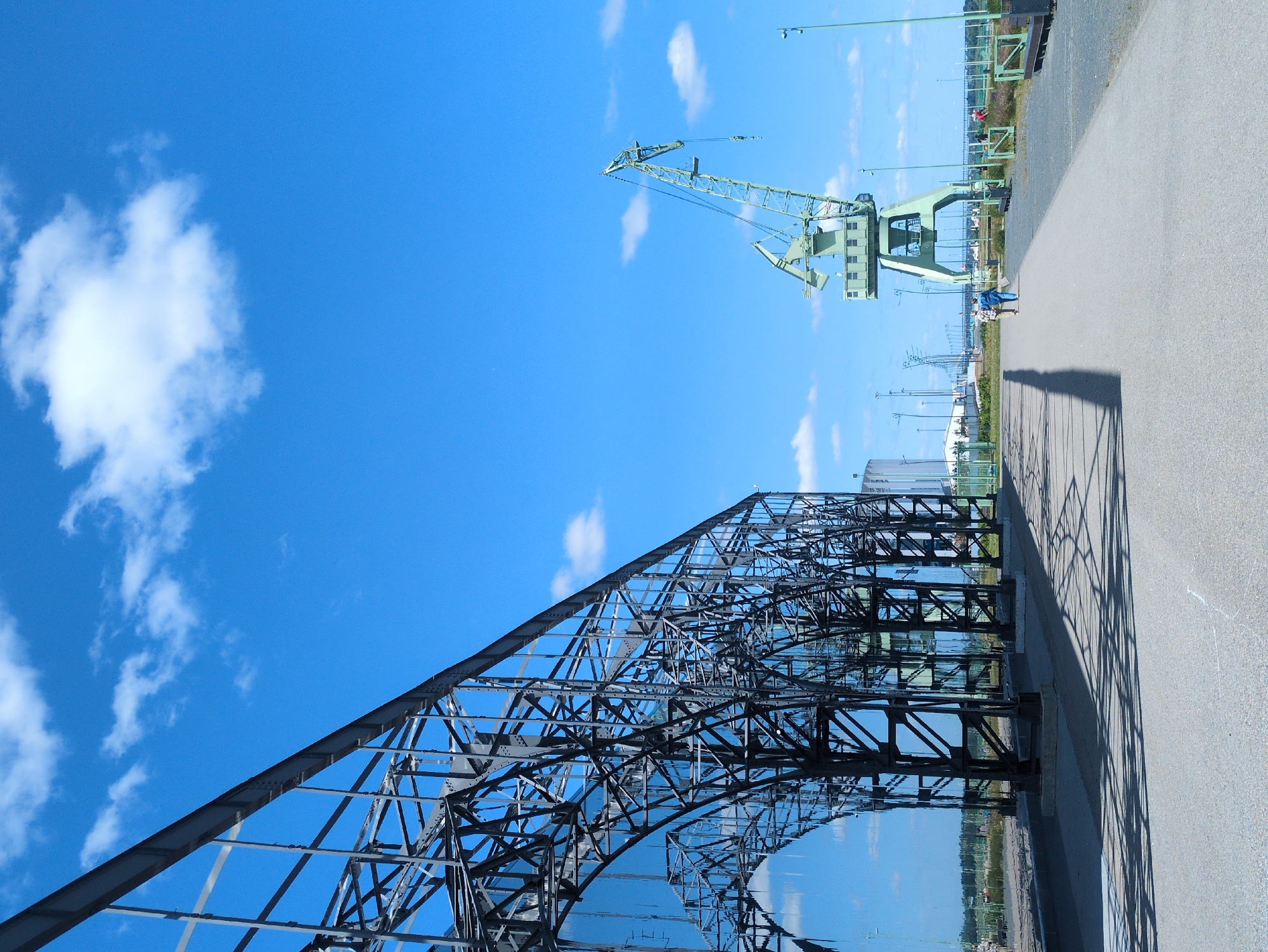This is a vertical, eye-level, full-shot photograph taken in an outdoor industrial area, possibly a port, in Nizhny Novgorod, Russia. On the left, a large, dark metal arched structure with a grid-like pattern takes up the left third of the frame, reflecting the blue sky with scattered white clouds. The structure casts a long, intricate shadow on the paved ground in front of it. To the right of this structure, a wide, light-grey paved path leads away from the viewer. Further down the path, two people are walking away from the camera. Beyond them and to the right, a tall, green industrial crane with a long boom stands prominently against the sky. The crane has a box-like structure at its base and a hook hanging from its boom. In the background, a row of tall poles with lights are visible, along with some low buildings and fencing. The overall environment appears to be a clear, sunny day.