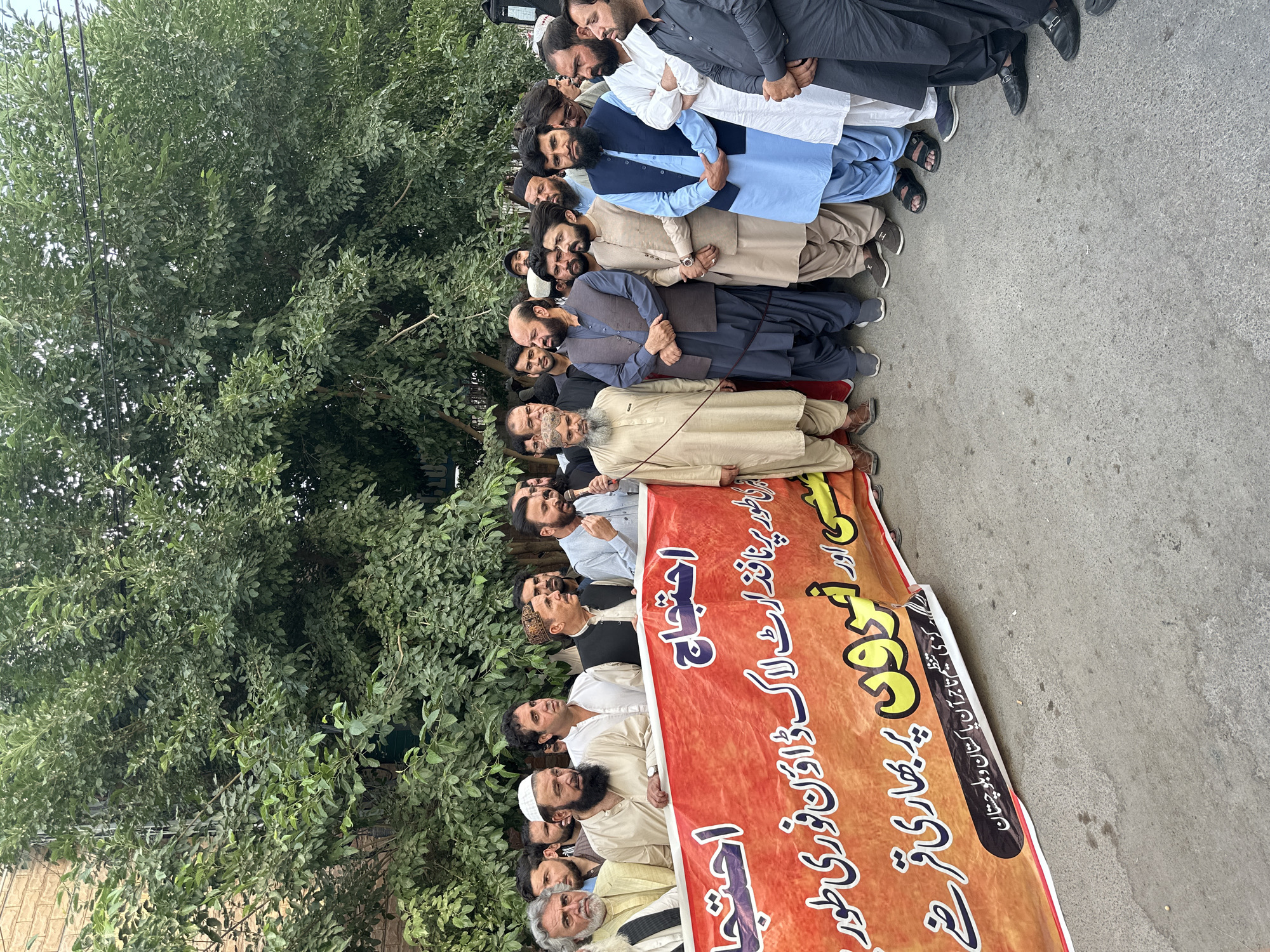 A group of men are standing in a row on a street in Quetta City Tehsil, Pakistan, holding a banner with writing in Urdu. The men are of various ages and are dressed in traditional Pakistani clothing. Some of the men are wearing turbans or prayer caps. The banner is red and gold and appears to be protesting something related to business or the economy. The background shows a tree and a brick building. The scene suggests a demonstration or public gathering.