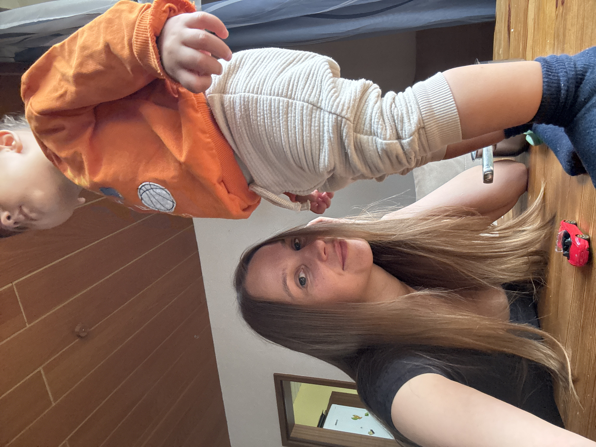 The image shows a selfie of a woman and a baby, likely taken indoors. The woman is in the foreground, with her head tilted back and her hair falling around her face. She is wearing a black t-shirt. The baby is in the background, facing away from the camera, wearing an orange hoodie and cream-colored pants. The baby's arm is visible, and it appears to be reaching out.
The setting appears to be a room with wooden paneling on the walls and a wooden table or surface in the foreground. A small red toy car is visible on the wooden surface near the woman's arm. The lighting suggests it is daytime, possibly indoors with natural light. The overall mood seems casual and intimate, given the selfie format and the presence of a baby.
There is no visible text in the image. The location context is Überherrn, Germany, but this information is not visually inferable from the image itself.