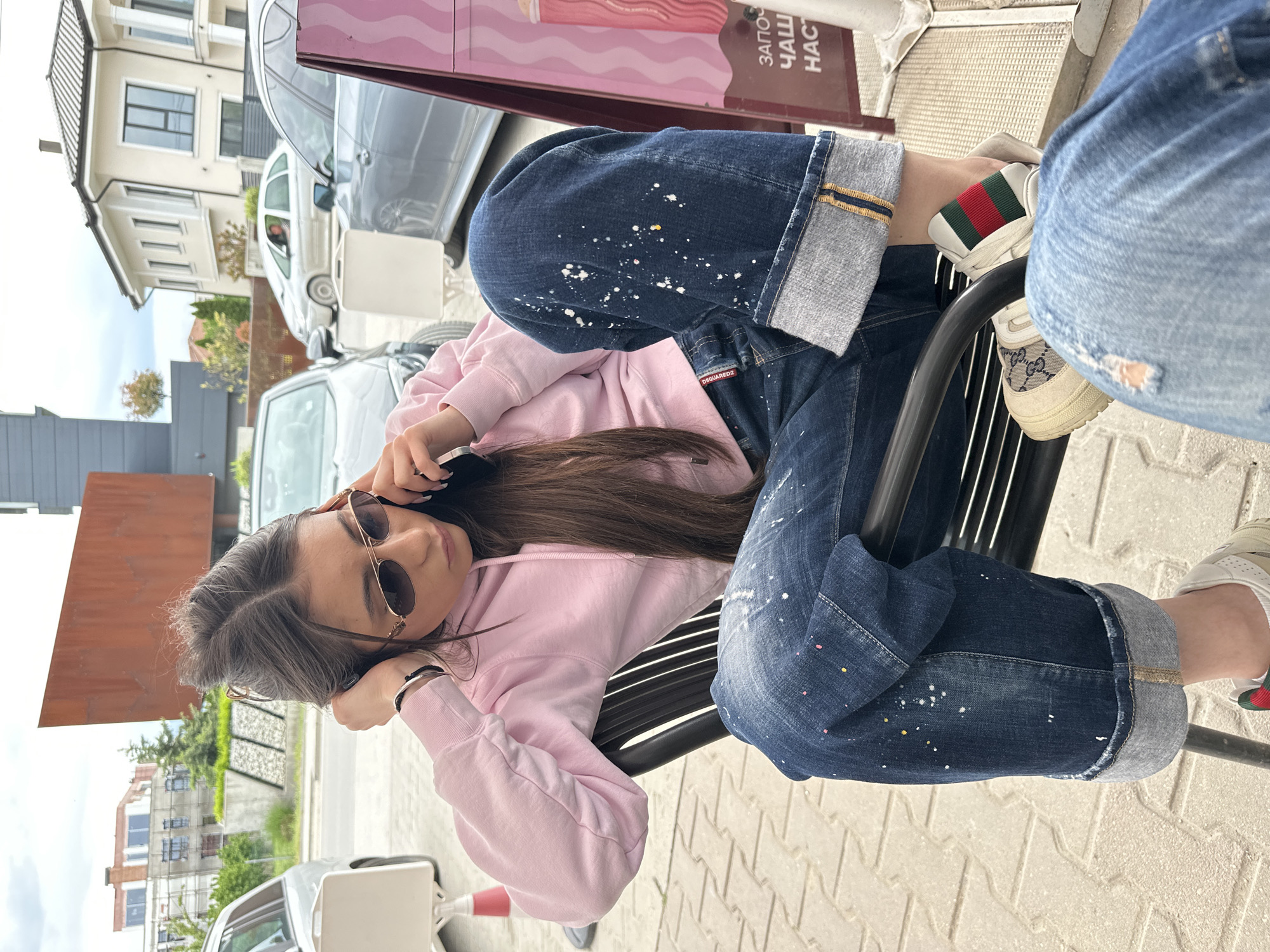 A young woman with long brown hair, wearing a pink hoodie, blue jeans with paint splatters, and sunglasses is sitting on a black metal bench outdoors. She is holding a black phone to her ear. She is wearing white and beige sneakers with green and red stripes. Another person is partially visible to her right, wearing ripped blue jeans. The location appears to be an urban environment, with buildings and parked cars visible in the background. Based on the text in the background, this location is Plovdiv, Bulgaria.