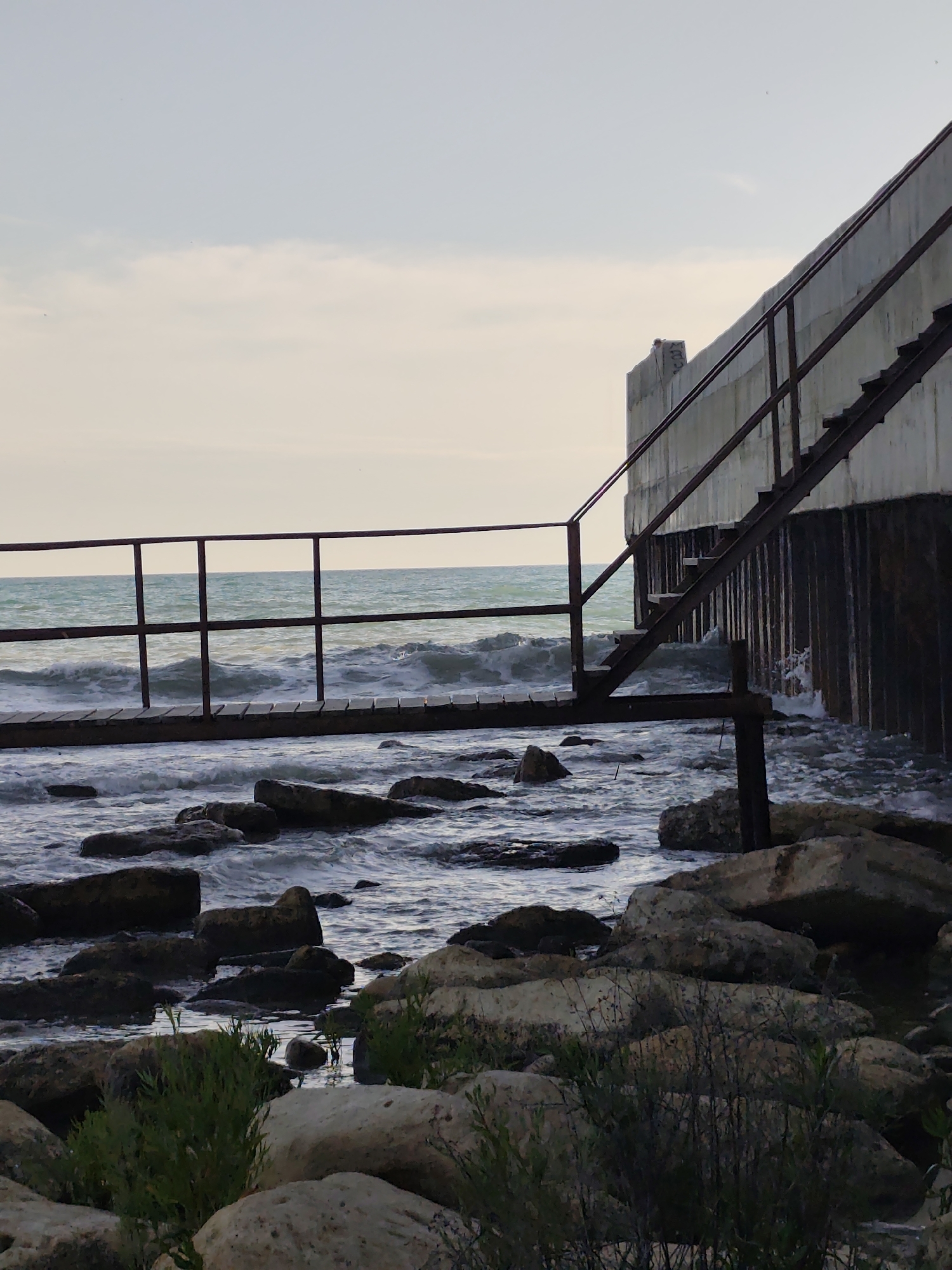 The image shows a concrete structure with metal stairs leading up its side, positioned next to a body of water. The water is choppy and appears to be the sea, with waves crashing against a rocky shore in the foreground. There is a metal walkway or bridge in front of the concrete structure, also with a metal railing. The sky is overcast, suggesting cloudy or hazy weather. The overall scene conveys a sense of industrial architecture meeting a natural, rugged coastline. The time of day appears to be either early morning or late afternoon due to the soft, diffused light. The location is likely a coastal area with man-made structures designed to withstand the elements. There are no people visible in the image. The concrete structure is imposing and utilitarian, possibly a part of a pier or a coastal defense. The rough texture of the rocks and the movement of the water add a dynamic element to the otherwise static architecture. The mood is somewhat somber and stark, highlighting the contrast between the man-made and natural environments. Some faint markings or graffiti are visible on the top part of the concrete structure.