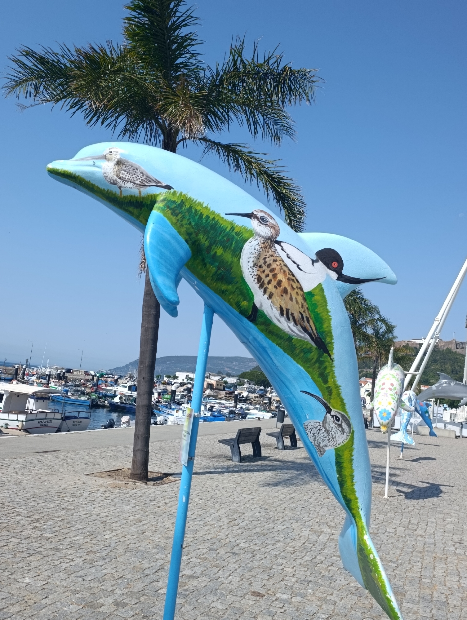 The image shows a large, blue dolphin sculpture painted with various seabirds and green grass. The sculpture is positioned outdoors on a cobblestone walkway. In the background, there is a harbor filled with boats, a palm tree, and buildings. The sky is clear and blue. The sculpture is located in Setúbal, Portugal, specifically along the waterfront promenade. There are no people visible in the image.