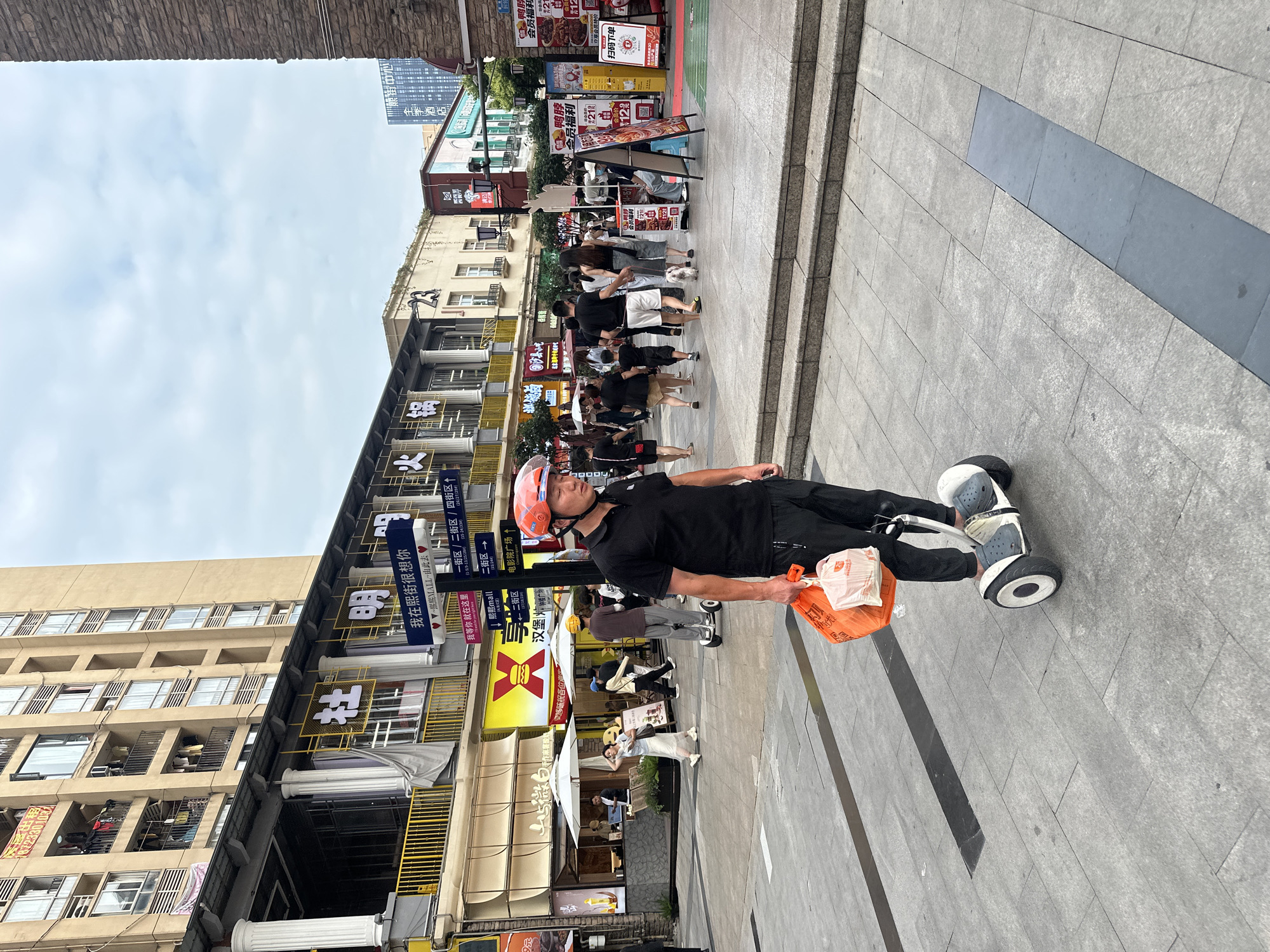 In an outdoor urban setting, a person wearing a red helmet and a black polo shirt rides a white self-balancing scooter. They are carrying two shopping bags, one orange and one white, in their right hand. The scooter has black wheels and grey footpads. The person is on a grey paved walkway with darker grey rectangular inlays. In the background, there are multi-story buildings with balconies and shops. Signage in Chinese characters is visible on the buildings, with some signs featuring yellow and red colors. Several other pedestrians are present in the background, some walking and others on scooters. The sky is overcast. The scene appears to be a bustling commercial street.