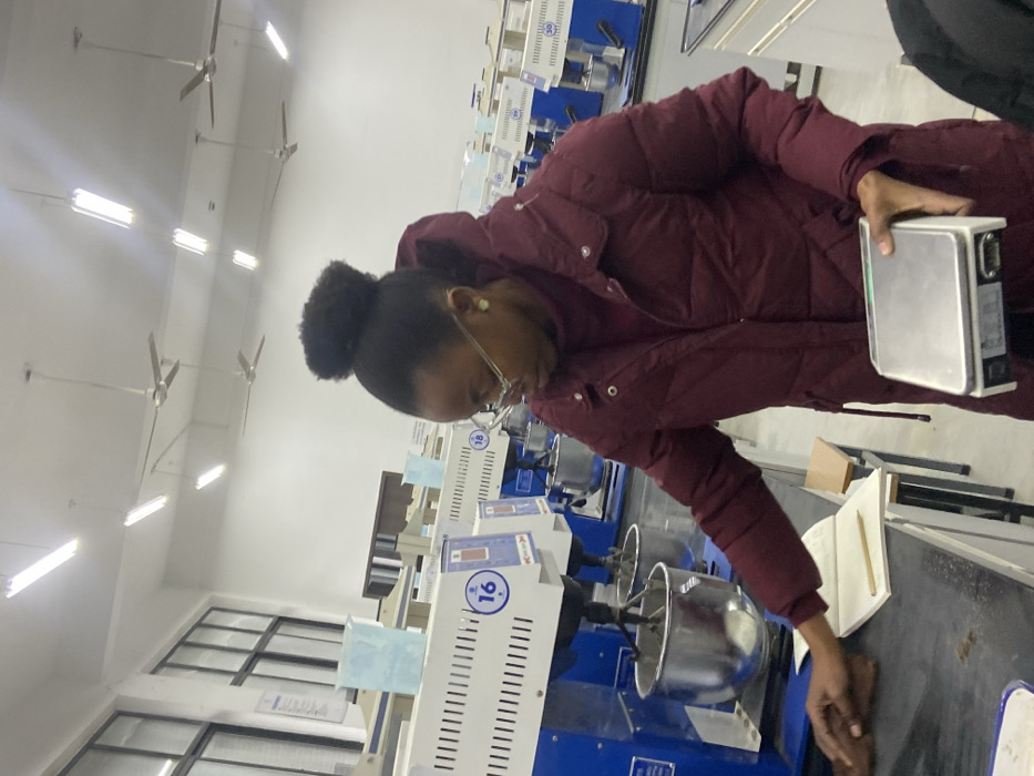 A woman in a maroon puffy jacket stands in a lab filled with identical blue and white machines. She holds a silver device in one hand and wipes the black countertop with the other. The machines each have a stainless steel mixing bowl. Fluorescent lights illuminate the room, and ceiling fans are visible in the background. A notebook and pencil are on the counter near the woman's hand.