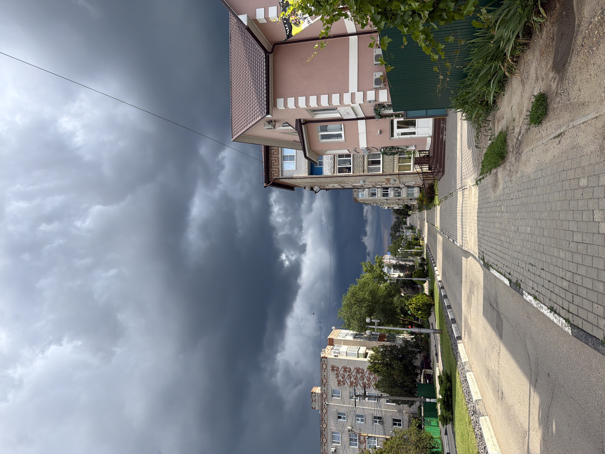The image captures a street scene in Leningradskaya, Russia, under a dramatic, dark, and cloudy sky that suggests an impending storm. The perspective is looking down a paved road with a pedestrian walkway to the right. Buildings line both sides of the street. On the left, a multi-story residential building with a patterned facade is visible. On the right, a lighter-colored, multi-story building with a pitched roof and decorative white trim stands out. Green fencing and lush foliage are present along the right side of the walkway. The street itself appears to be in decent condition, with some wear and tear visible on the asphalt. No people are clearly visible in the image, and no specific actions are depicted. The overall environment suggests a quiet urban setting during potentially inclement weather.