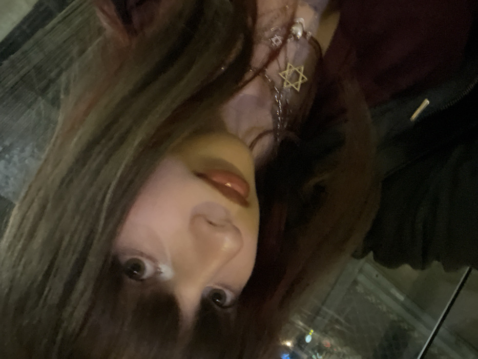 A person with dark brown hair framing their face is tilted to the side. They have fair skin, dark eyes, and glossy lips. A silver necklace with a Star of David pendant is visible. The background appears to be a window reflecting city lights at night. The person is wearing a dark-colored jacket or shirt.