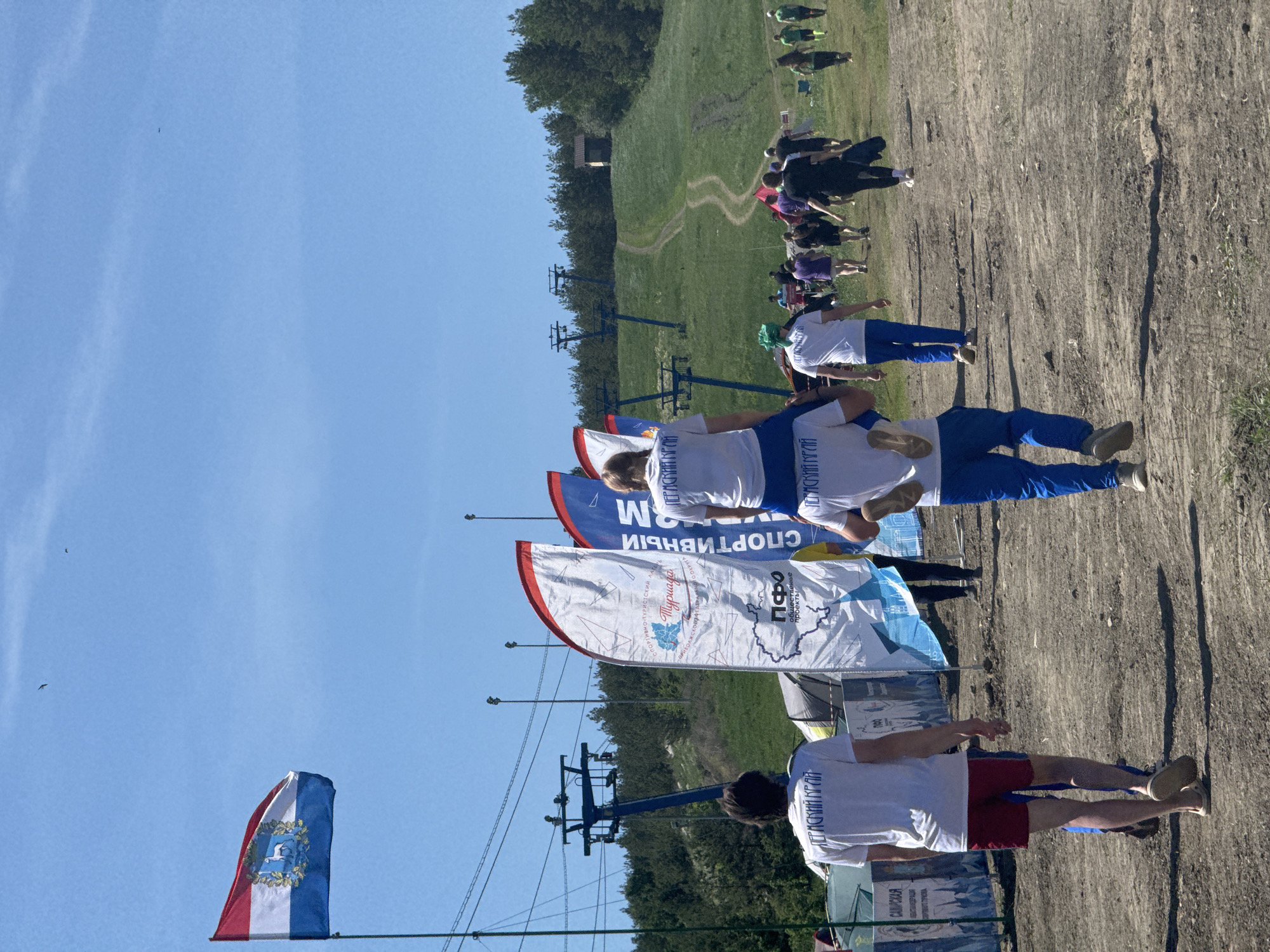 The image captures a scene from Khvalynsk, Russia. A Russian flag flutters on a pole in the foreground, with banners adorned with Russian text and symbols lining the area. People in white shirts and blue pants are visible, likely participants or organizers of an event. A ski lift structure stretches across the frame, ascending towards a grassy hill in the background where additional people are gathered. The environment suggests an outdoor recreational area, possibly a ski resort during the off-season. The ground appears to be dirt or gravel, indicating a pathway or open space. The sky is clear and blue, suggesting a sunny day.
