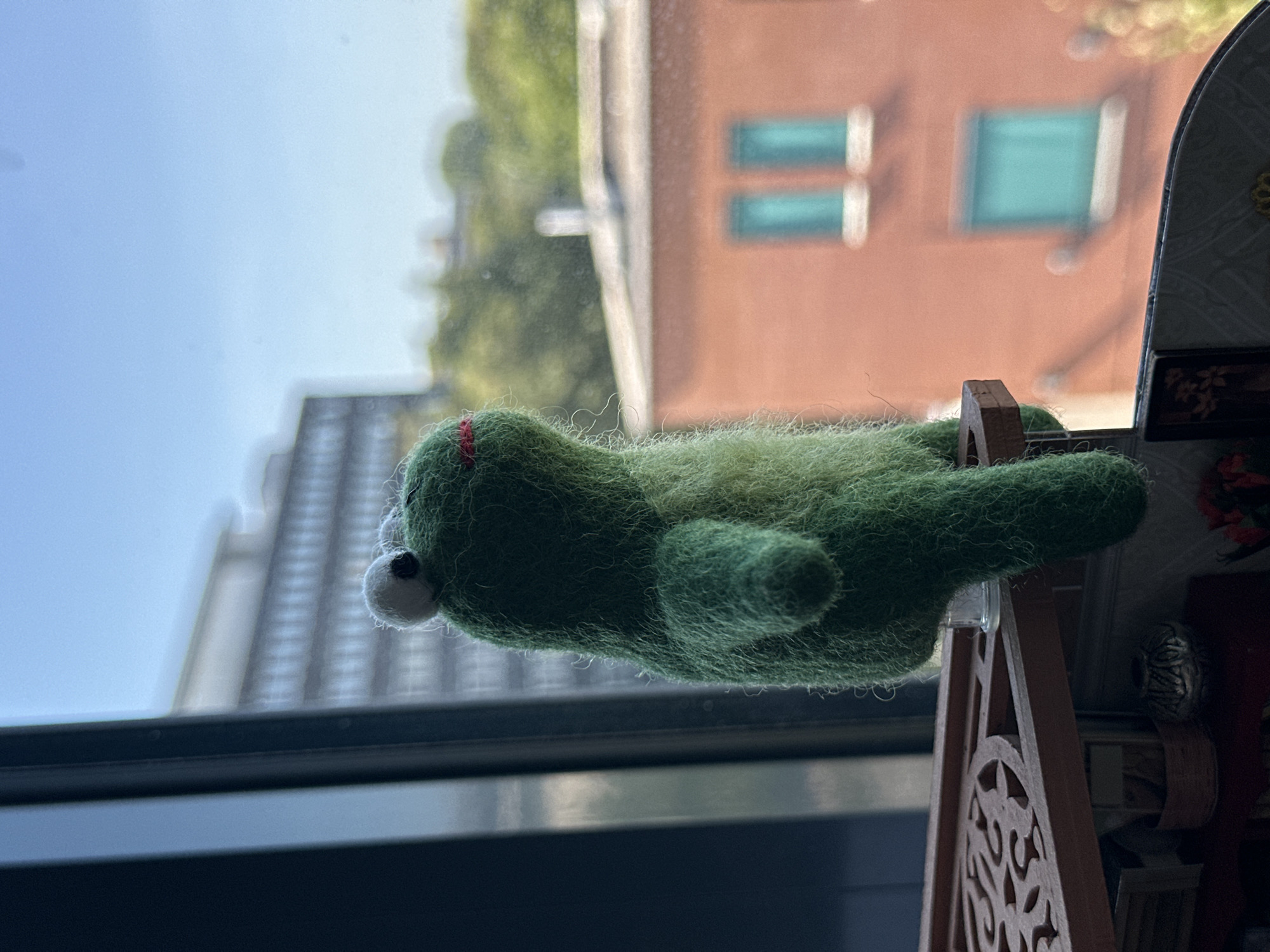 A needle-felted frog figurine is positioned on a small wooden ledge, looking out a window. The figurine is green with white google eyes and a small red stitched mouth. Behind the frog, through the window, is a blurry cityscape with buildings and trees. The ledge the frog is on is part of a miniature diorama, with other small objects and furniture visible below. The overall lighting suggests it is daytime, with sunlight filtering in from the window.