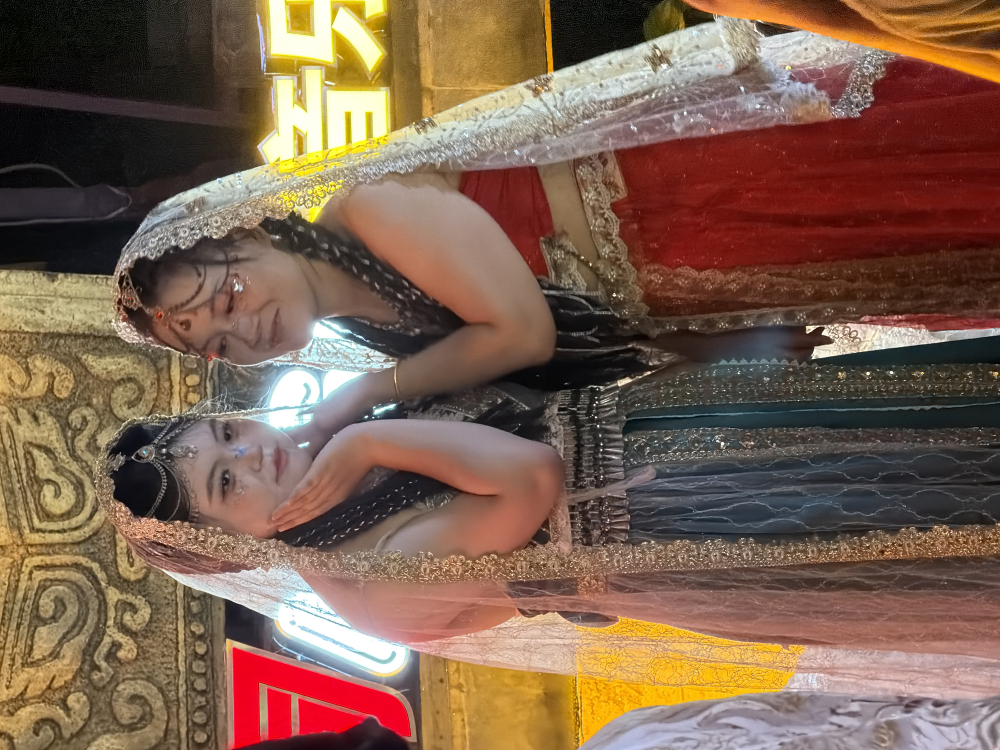 Two young women, dressed in elaborate traditional Indian attire with veils and ornate jewelry, are posing for a photo in what appears to be an outdoor night setting. The woman on the left wears a dark green and grey dress with gold trim, while the woman on the right is in a red dress with similar gold accents. Both have intricate makeup, including glitter on their faces. Behind them, a brightly lit sign with Chinese characters is visible, and the background suggests an urban environment with some architectural features. The scene is likely within the Yuzhong District of Chongqing, China, which is known for its vibrant nightlife and illuminated cityscape, often featuring elaborate decorations for festivals or events. The lighting suggests artificial light sources, common in urban night scenes.