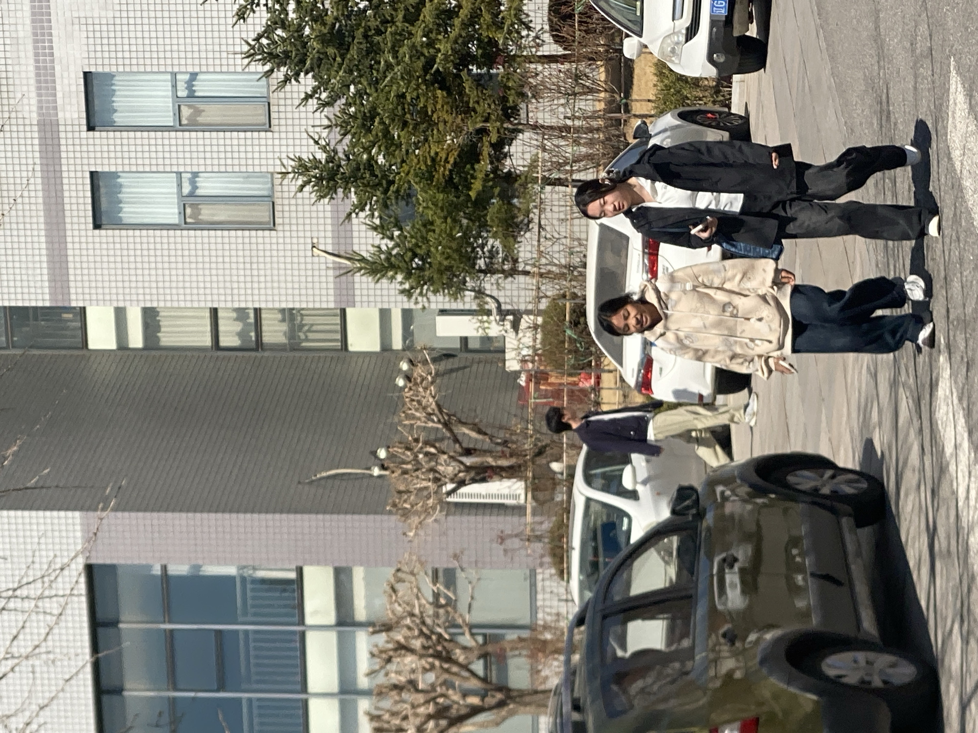 The image depicts an outdoor street scene under clear skies in Zhongtun, China. In the foreground, two young women walk left-to-right across an asphalt surface with faint white road markings. The woman on the left wears a light-colored hooded sweatshirt and dark trousers, holding a white object in her left hand. The woman on the right wears a black jacket over a white shirt, dark trousers, white shoes, and carries a blue bag on her left shoulder while holding a white smartphone in her right hand.
Behind them, a line of parked vehicles is visible. From left to right, these include a dark olive-green crossover SUV, a white sedan, and the front portion of a white SUV. Further in the mid-ground, a young man in a dark jacket and light trousers walks right-to-left between parked cars.
The background features multiple multi-story buildings under direct sunlight. The leftmost building has a light-colored tiled facade with large, reflective windows. The building on the right also has a light-colored tiled facade with smaller, vertically oriented windows, some showing interior curtains. Leafless, pruned deciduous trees are visible in the mid-ground, alongside several evergreen conifer trees situated in front of the rightmost building. Shadows are cast by the people and vehicles, indicating strong sunlight from the left side of the frame.