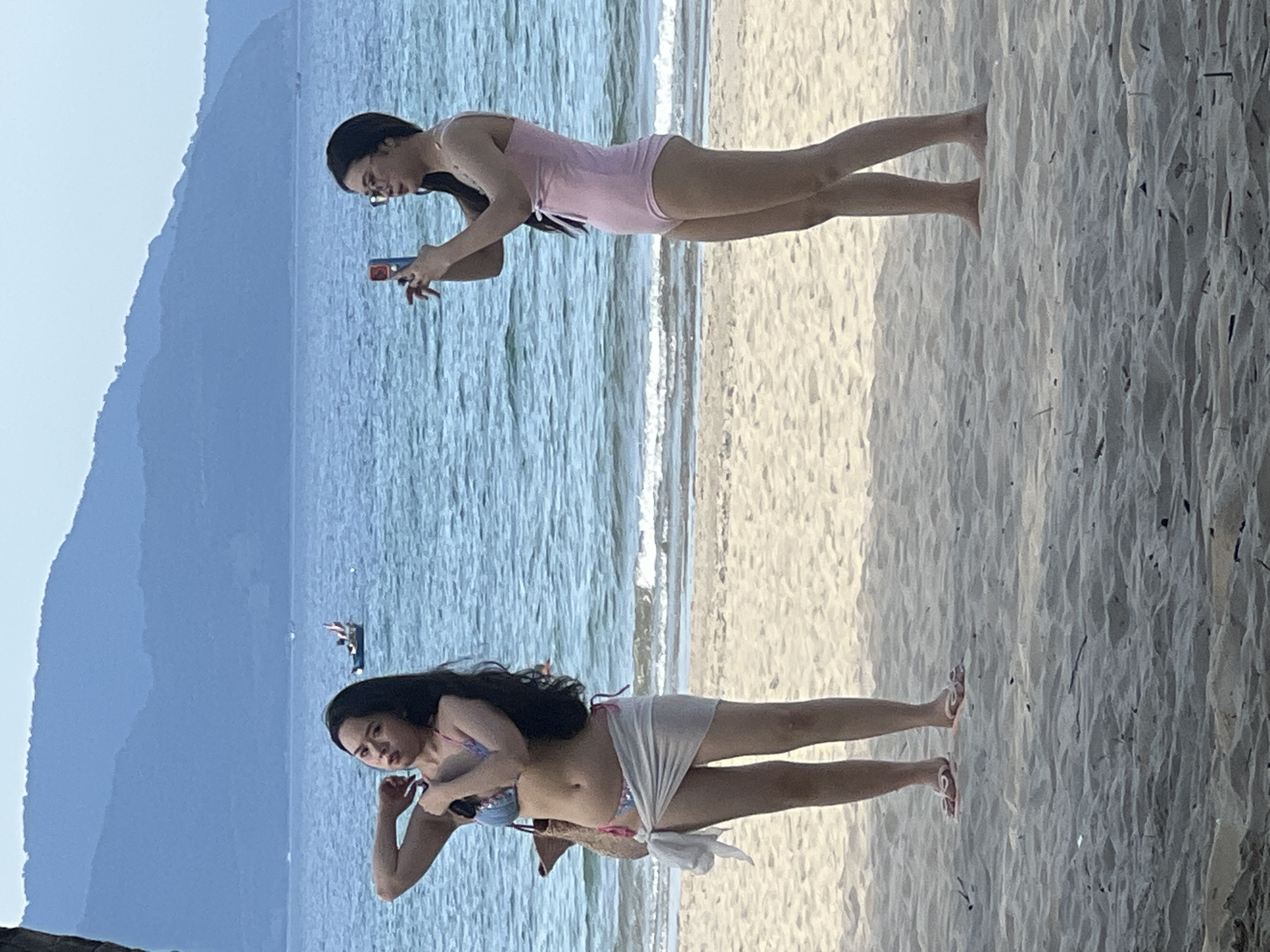 Two young women are standing on a sandy beach in Đà Nẵng, Vietnam, with the ocean and mountains in the background. The woman on the left is wearing a patterned bikini with a white sarong tied around her waist. She has her arms raised and is posing. The woman on the right is wearing a pink one-piece swimsuit and glasses. She is holding a smartphone and appears to be taking a photo or video. The water is blue and calm, with small waves breaking on the shore. The mountains in the distance are covered in lush green vegetation. The sky is overcast.