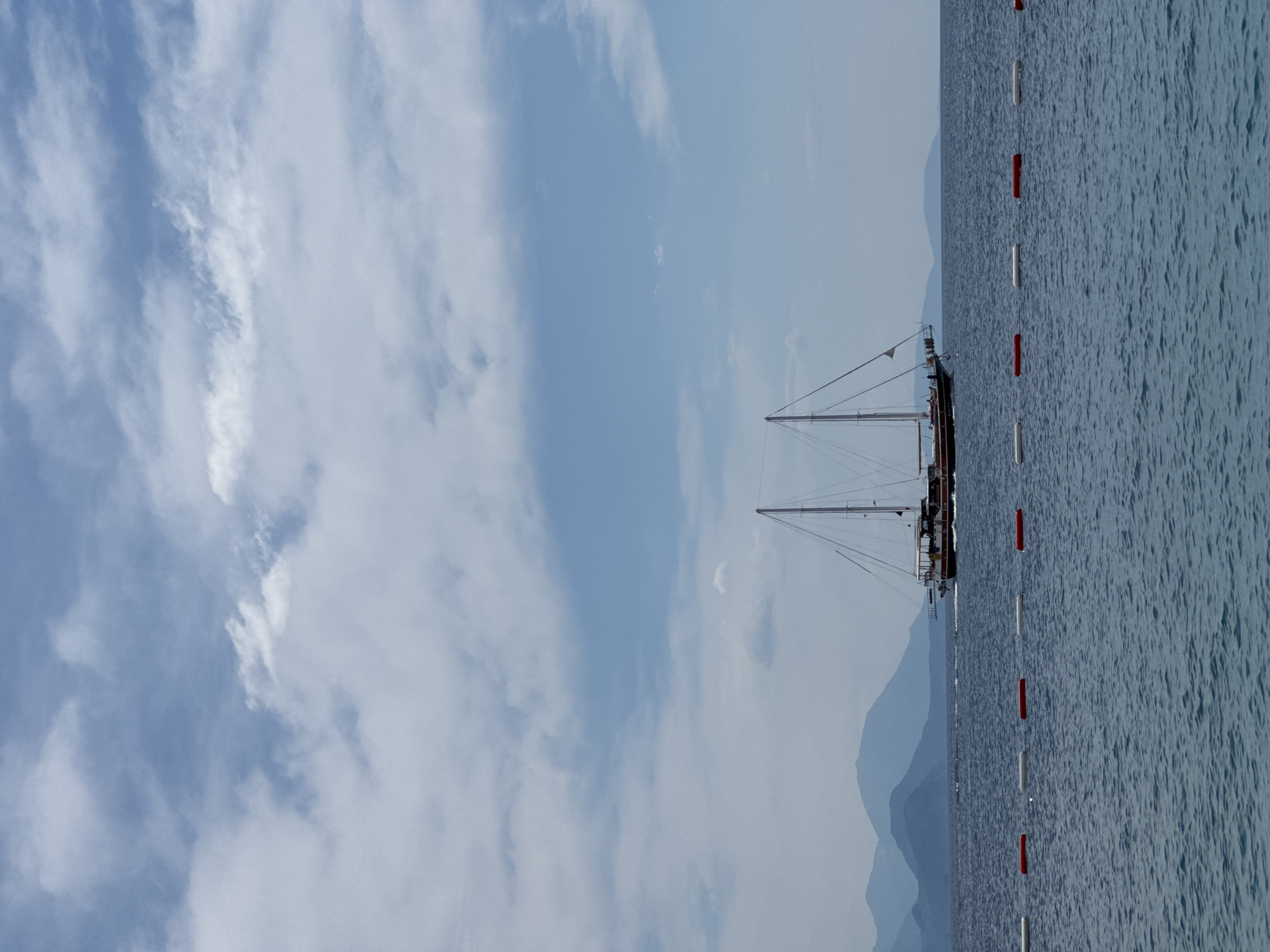 The image shows a seaside scene, possibly in Prijevor, Montenegro. The sky is blue with scattered clouds. A two-masted sailboat is anchored near a line of red and white buoys in the sea. Mountains can be seen in the background. The sea is calm with a slight ripple effect.