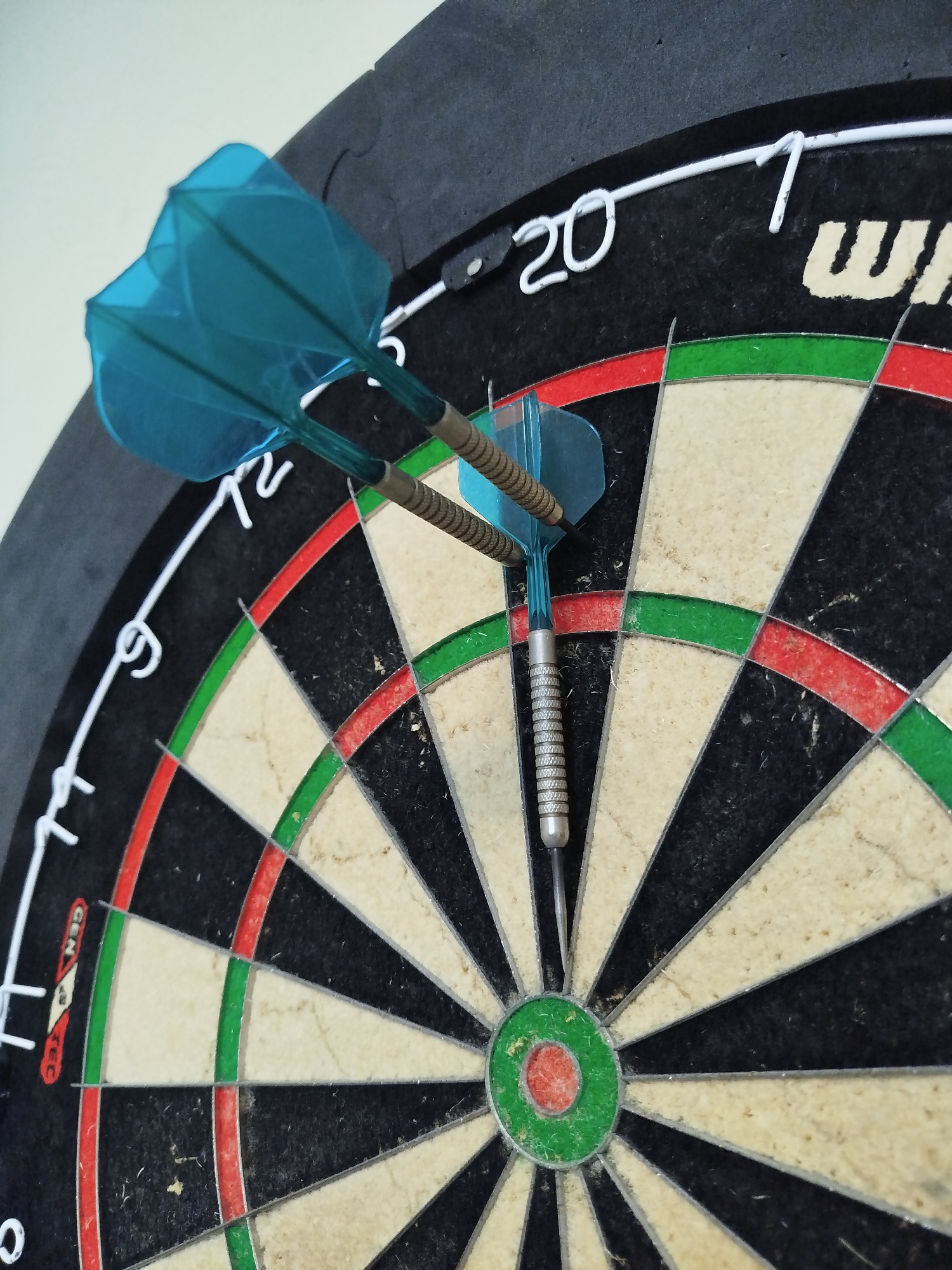 A close-up image shows a dartboard with three blue darts embedded in it. Two darts are clustered near the number 12 on the board, while the third dart is lodged in the bullseye. The dartboard is mounted on a wall and shows signs of use, with some wear and tear visible on the surface. The numbers and rings on the board are clearly discernible, and the overall image is well-lit. There is no indication of Varna, Bulgaria.