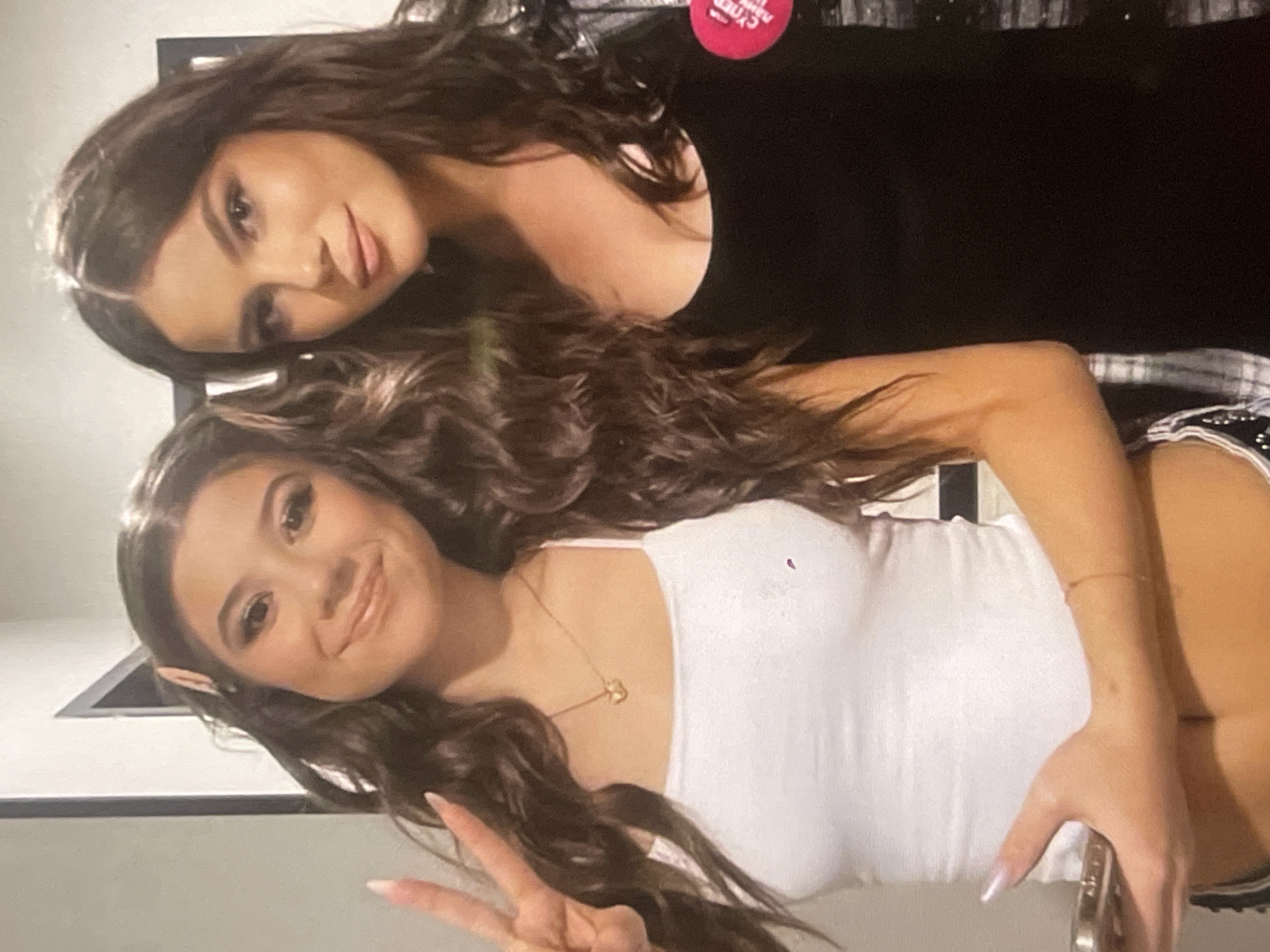 The image captures two young women posing closely together indoors in Poltava, Ukraine. Both have long, dark, wavy hair and wear full makeup.
The woman positioned slightly behind and to the left in the frame wears a black top. She leans her head gently on the other woman's shoulder, looking towards the camera with a soft, friendly smile. A round red badge with partially visible white text, appearing to read "CYBER" (mirrored in the image), is pinned to her top.
The woman in the foreground, positioned to the right, wears a white spaghetti strap top. She smiles broadly at the camera, making a peace sign with her right hand, which displays long, light-colored fingernails. A delicate gold necklace with a small pendant is visible around her neck. A distinct feature is the pair of prosthetic elf ears attached to her natural ears, suggesting a costume or playful accessory.
The background behind the woman in black is dark and indistinct, while the background behind the woman in white is a lighter wall with a dark, angular architectural element. The overall mood is cheerful and friendly, indicating a social gathering or event, possibly in the evening given the indoor setting and lighting.