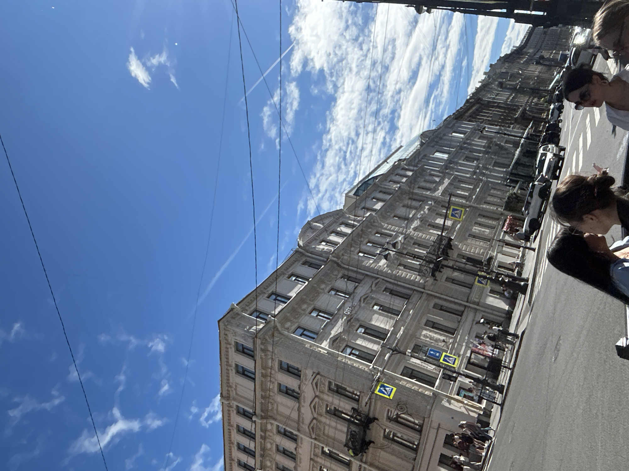 The image captures a city street scene on a sunny day. In the foreground, two women are visible from the chest up, with their backs to the camera, looking towards the street. The woman on the left has dark hair, and the woman on the right has brown hair styled in a bun and is wearing sunglasses.
The middle ground of the image shows a wide street with a crosswalk. Several cars are present on the street, including a white SUV and a white sedan. Pedestrians are walking on the sidewalk to the left of the street, in front of a large, ornate building. This building, which appears to be a hotel based on the word "ОТЕЛЬ" visible on its facade, has multiple stories with many windows and balconies. Traffic signs, including yellow pedestrian crossing signs, are posted along the street.
The background features more of the same architectural style of buildings lining the street, stretching into the distance. The sky is a bright blue with scattered white clouds and visible contrails from airplanes. Overhead power lines crisscross the sky. The overall impression is of a vibrant, sunny day in an urban environment.