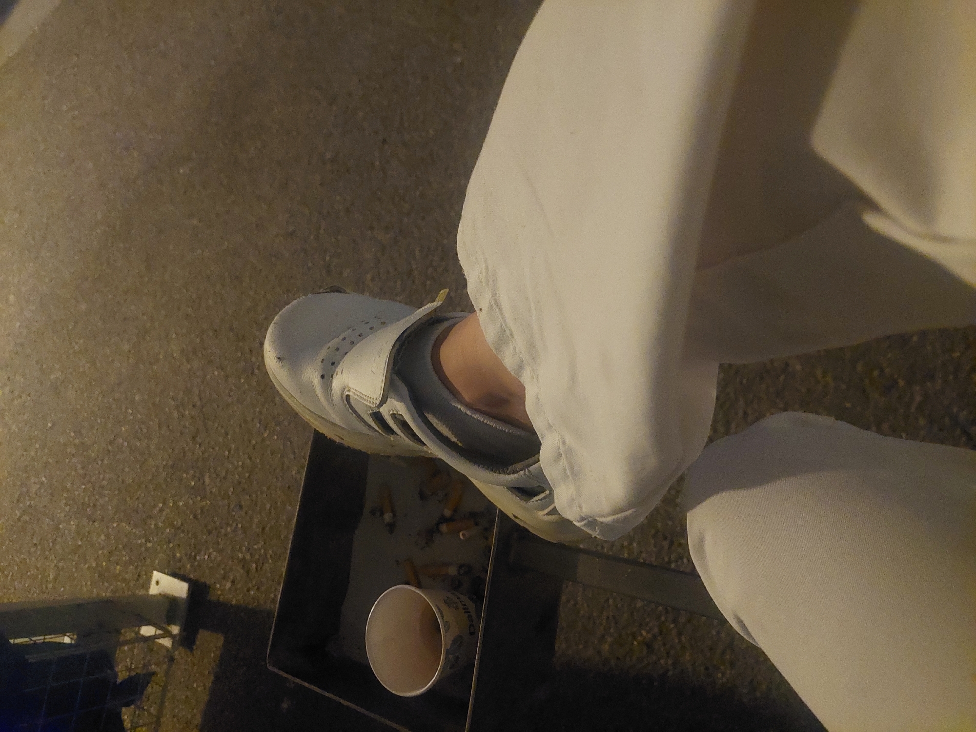 A high-angle, close-up shot shows a person's leg and foot in Altdorf, Germany, positioned next to an ashtray. The person wears white pants and a light gray athletic shoe with velcro straps. The ashtray is a dark gray metal container filled with cigarette butts. Beside the cigarette butts is a partially empty paper cup. The ground around the ashtray appears to be paved. A small section of a blue metal structure is visible in the lower left corner. The scene is dimly lit, possibly at night.