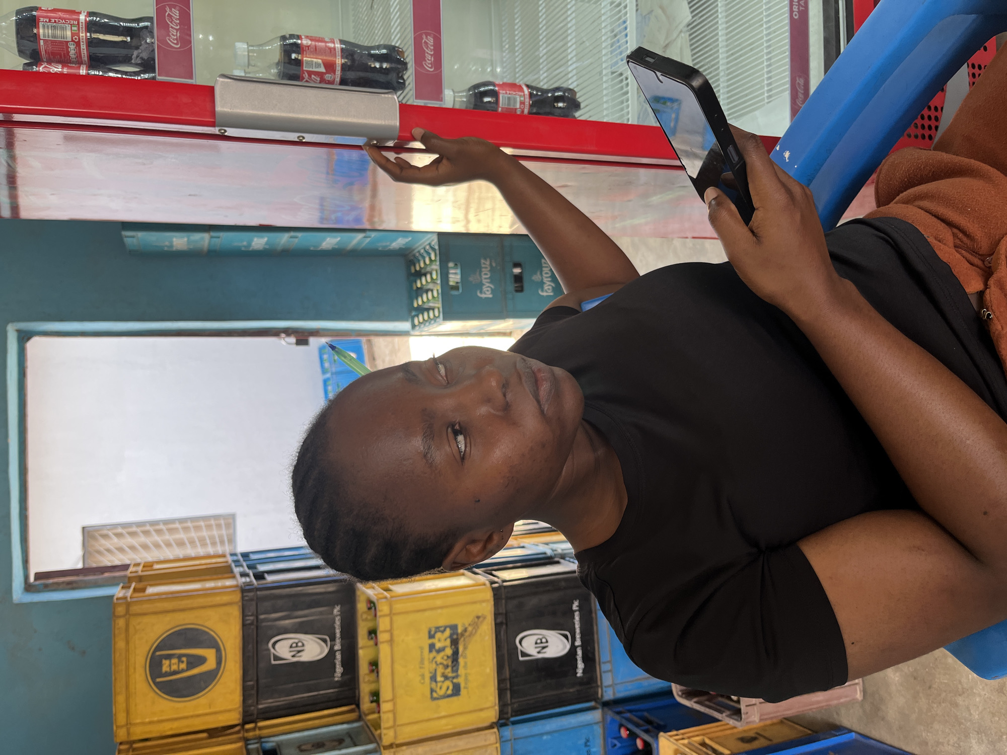 A woman is seen looking at a smartphone in what appears to be a small retail space in Jos, Nigeria. She is seated on a blue plastic chair and is wearing a black t-shirt. The location seems to be a shop or small store where beverages are sold, as evidenced by the Coca-Cola refrigerator in the background and stacks of crates bearing the logos of Nigerian Breweries and STAR beer, all located to the left. The crates are yellow, blue and black. The refrigerator contains Coca-Cola bottles and also has signage. The lighting is relatively dim, suggesting an interior setting.