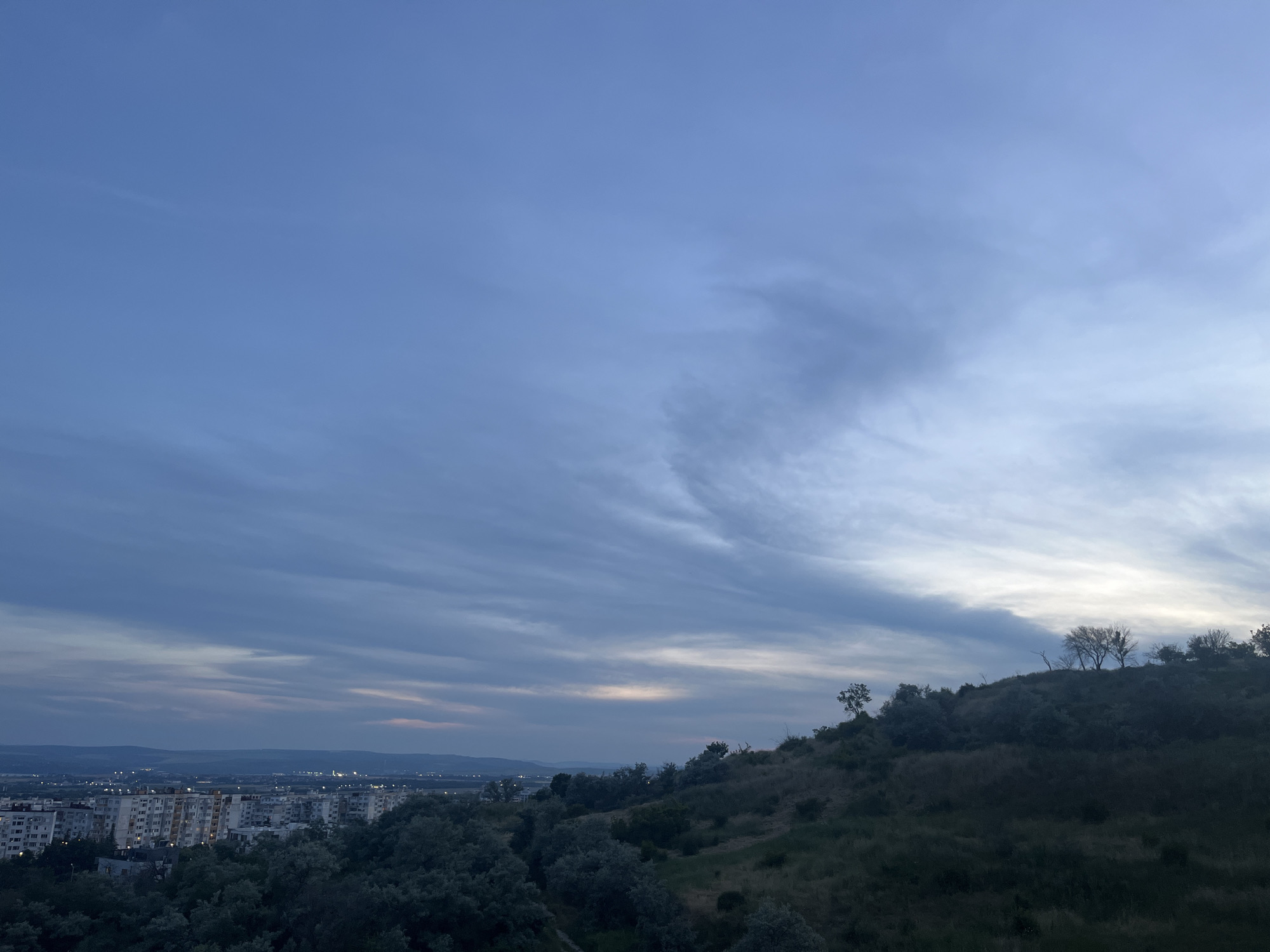 This is a high-angle, full-frame shot of a cityscape at dusk, taken from a hilltop overlooking the city of Varna, Bulgaria. The sky is a deep blue with scattered clouds, tinged with pink and yellow near the horizon, indicating the setting sun. In the foreground, a grassy hill slopes down towards the city, covered in dark green trees. The city itself is spread out below, featuring numerous apartment buildings, some with lit windows, and further in the distance, rows of houses and industrial structures. The overall mood is serene and quiet, with no people visible in the frame. The image captures a typical urban landscape at the end of the day.