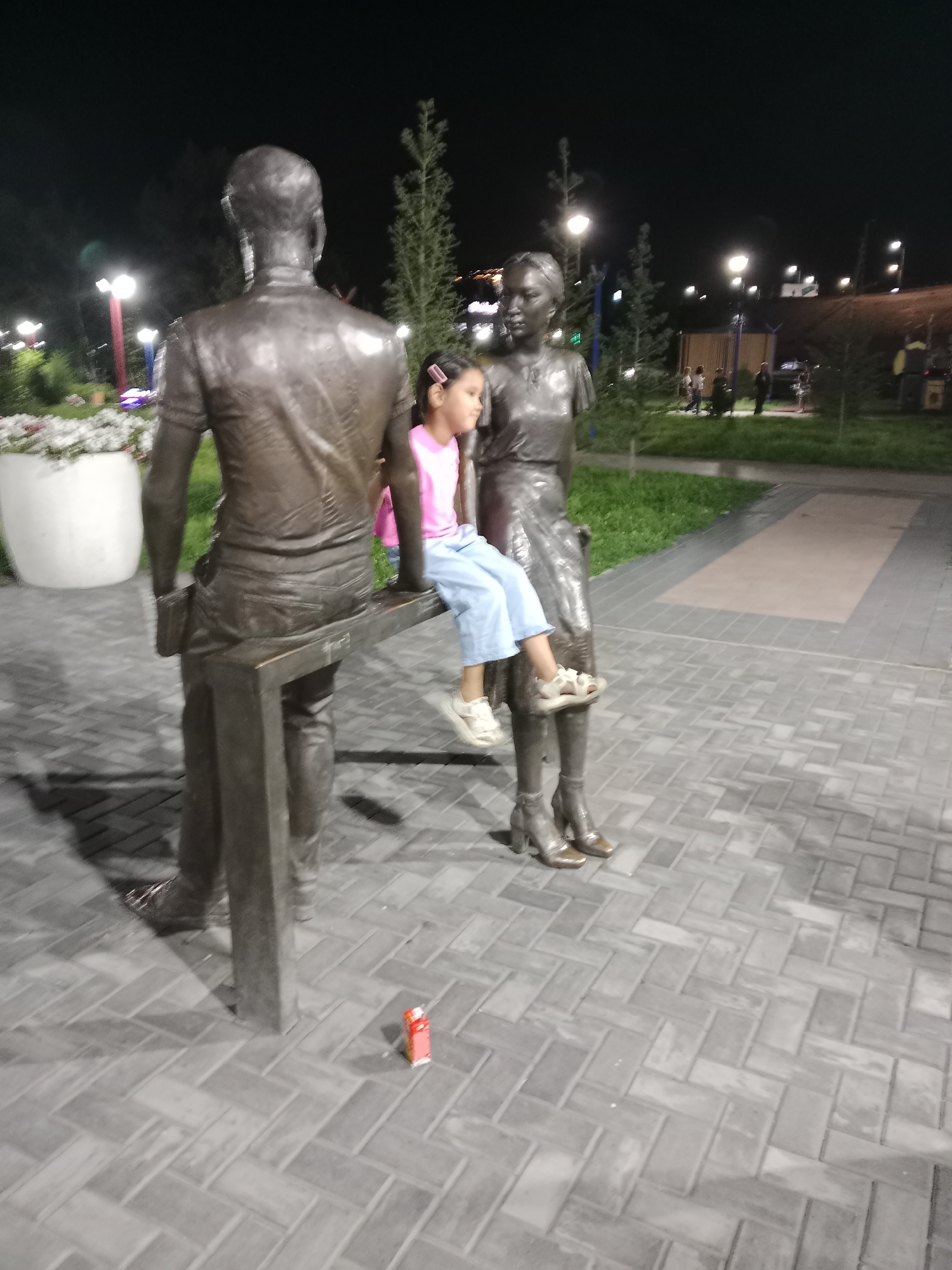 A nighttime outdoor scene depicts a bronze sculpture of a man and a woman seated on a bench. A young girl, wearing a pink shirt and blue pants, is seated on the bench between the two figures of the sculpture. The girl is looking to her right. The sculpture is located in a park or public space with paved ground and some greenery and trees in the background. Streetlights illuminate the area. A small red juice box lies on the ground near the sculpture's base. The image was taken in the city of Semey, Kazakhstan.