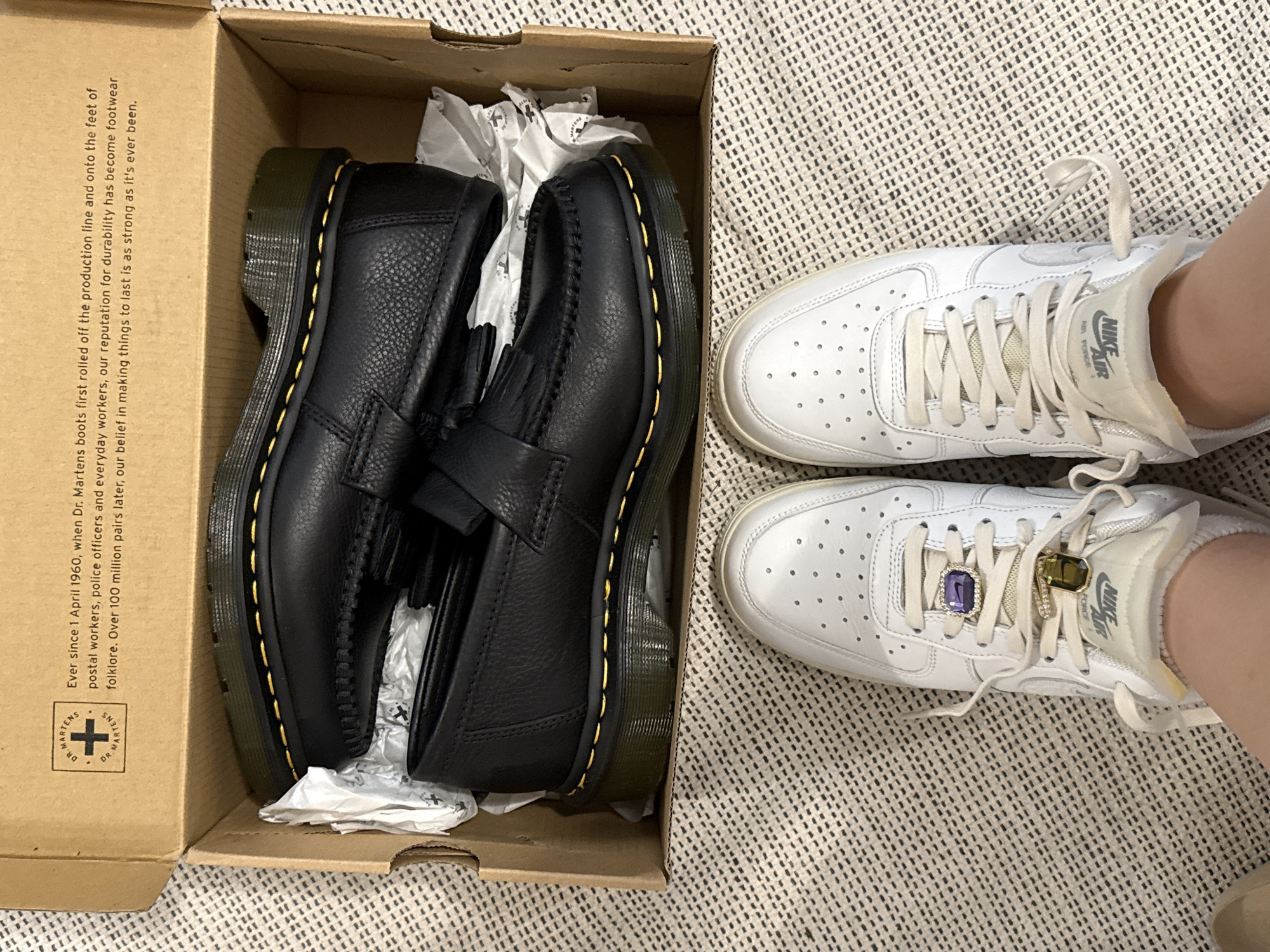 This indoor overhead shot features two pairs of shoes on a light-colored, textured rug. On the left, an open brown cardboard Dr. Martens shoe box contains a new pair of black leather loafers. The loafers have a pebbled texture, distinctive yellow stitching around the sole, and fringed tassels on the vamp. White crumpled tissue paper is visible inside the box around the shoes.
On the right, a person's feet are visible, wearing white Nike Air Force 1 sneakers. The right sneaker is adorned with two rectangular, jewel-like charms on its laces: one purple and one olive green. The sneakers appear to be slightly used but well-maintained.
Visible text on the inside flap of the Dr. Martens box reads: "Ever since 1 April 1960, when Dr. Martens boots first rolled off the production line and onto the feet of postal workers, police officers and everyday workers, our reputation for durability has become footwear folklore. Over 100 million pairs later, our belief in making things to last is as strong as it's ever been." Below this is the "DR. MARTENS" logo. The tongue of the Nike Air Force 1 sneaker displays "NIKE AIR FORCE 1" branding. The scene suggests the unboxing of new shoes or a comparison of different footwear.
