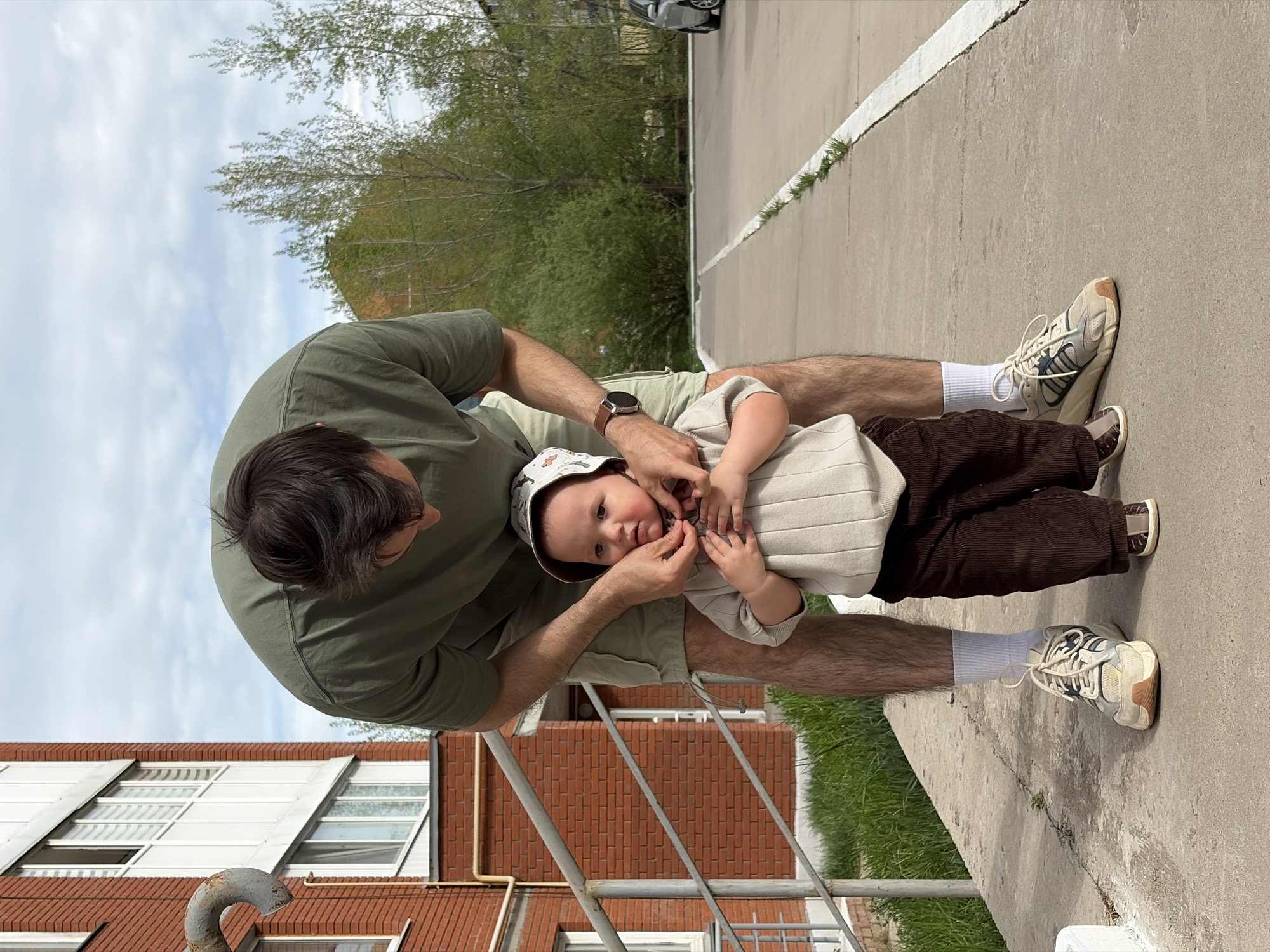 A medium shot of a man and a toddler outside a brick building. The man, dressed in an olive-green t-shirt and khaki shorts, is crouched down, adjusting the collar of the toddler's outfit. The toddler, wearing a hat, a beige striped top, and brown corduroy pants, looks at the camera. Both are situated on a concrete surface next to a grassy area and a railing. In the background, there are trees and a glimpse of the sky. Part of a building with windows is visible on the left. The location is Chaikovsky, Russia.